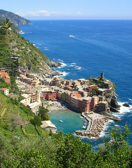 Panoramic view of Vernazza village with colorful buildings, rocky coastline, and blue sea, surrounded by green hills and terraced vineyards