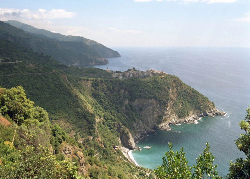 Coastal landscape with steep green cliffs, turquoise sea, and village structures on a hillside under a partly cloudy sky.