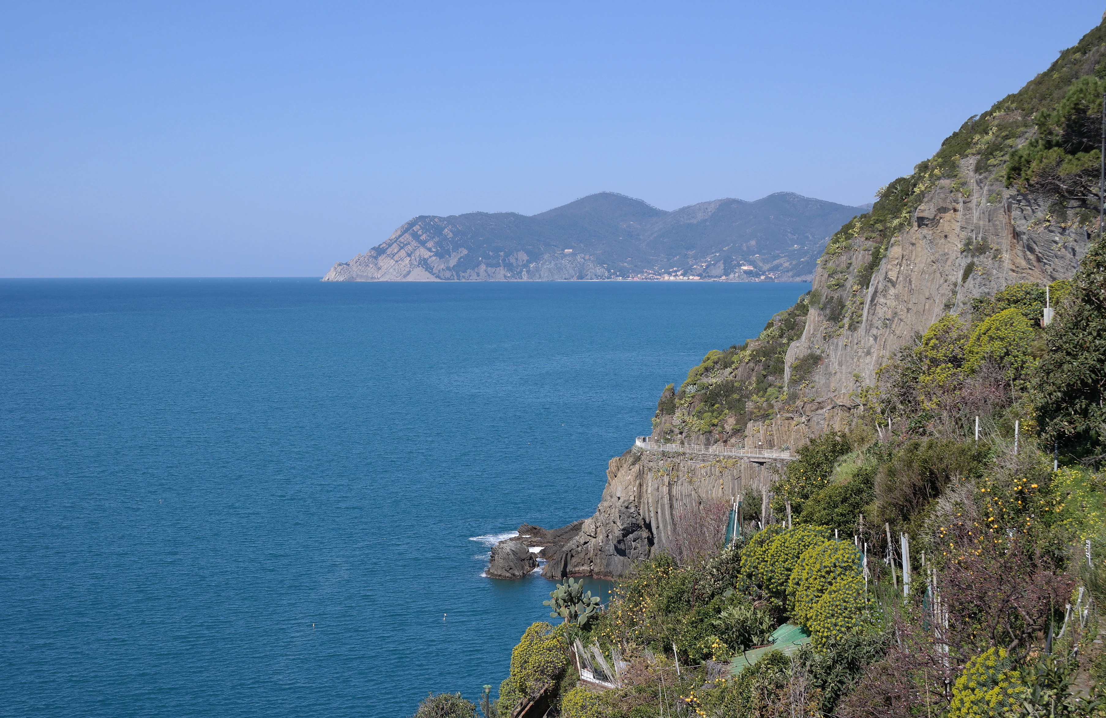 Coastal panorama showing blue sea, rocky cliffs, lush vegetation, and distant mountains under clear blue sky