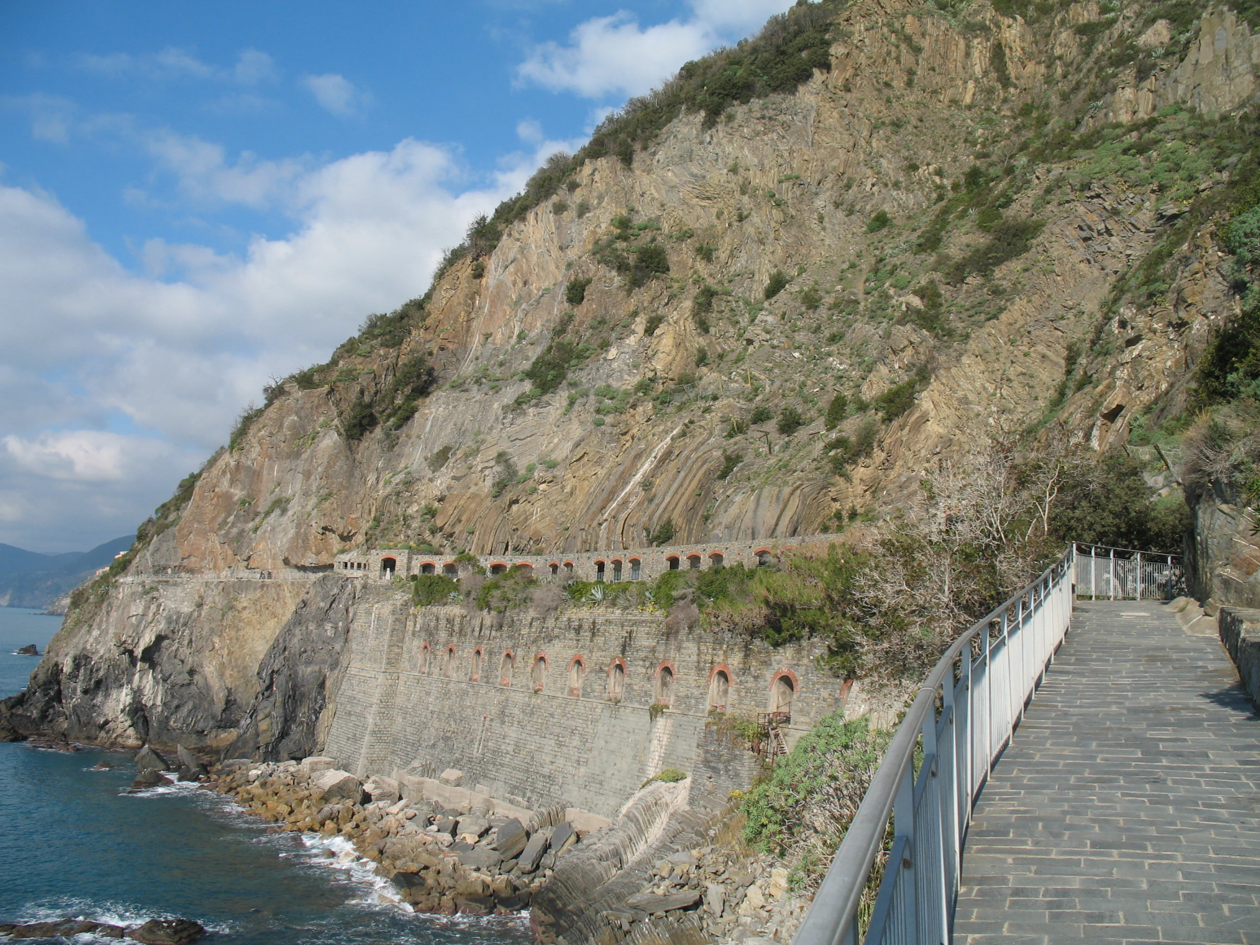 Stone pathway with metal railing alongside rocky cliffs overlooking the sea