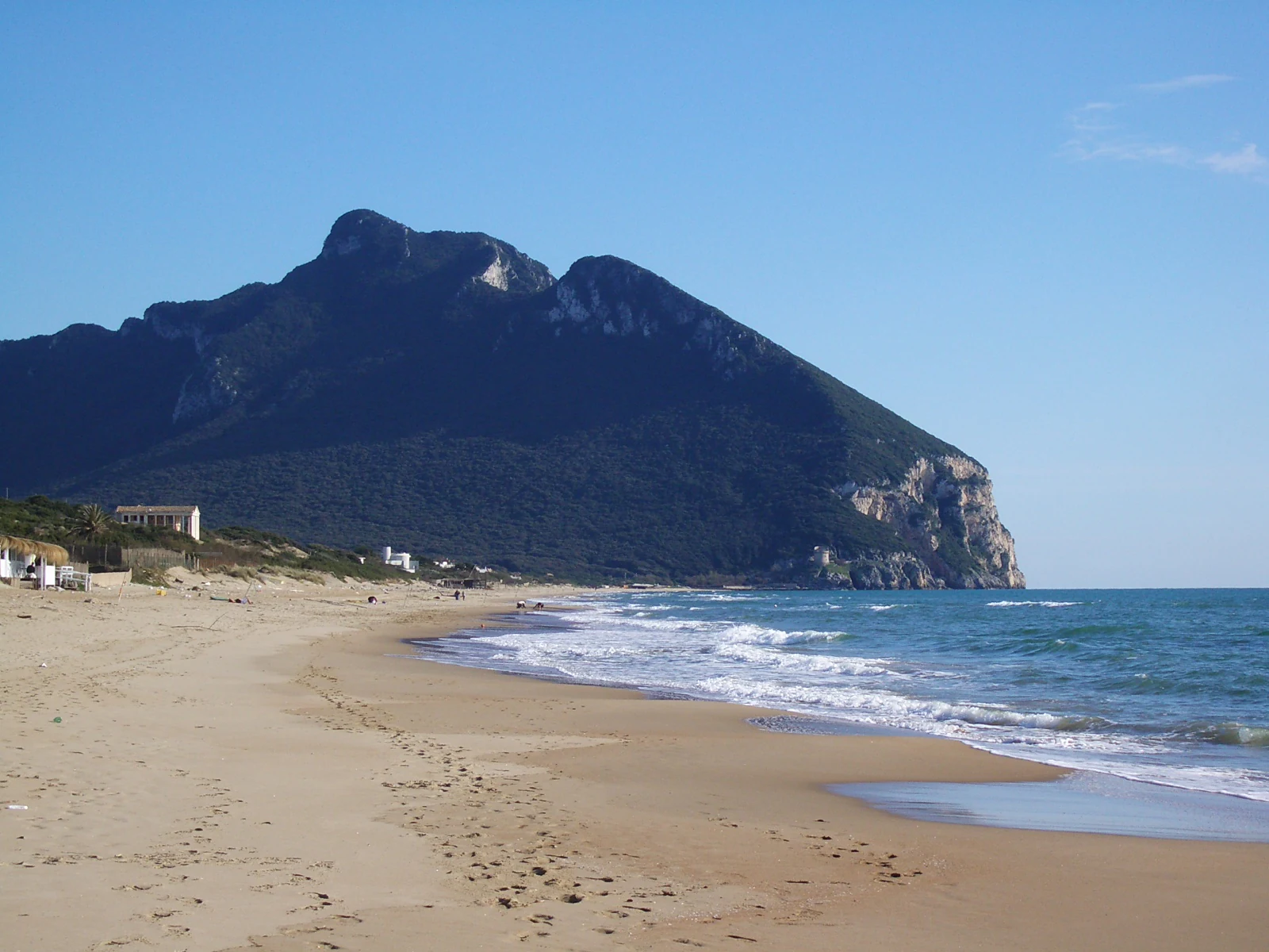Sandy beach with footprints, gentle waves, a mountain with vegetation, and clear blue sky