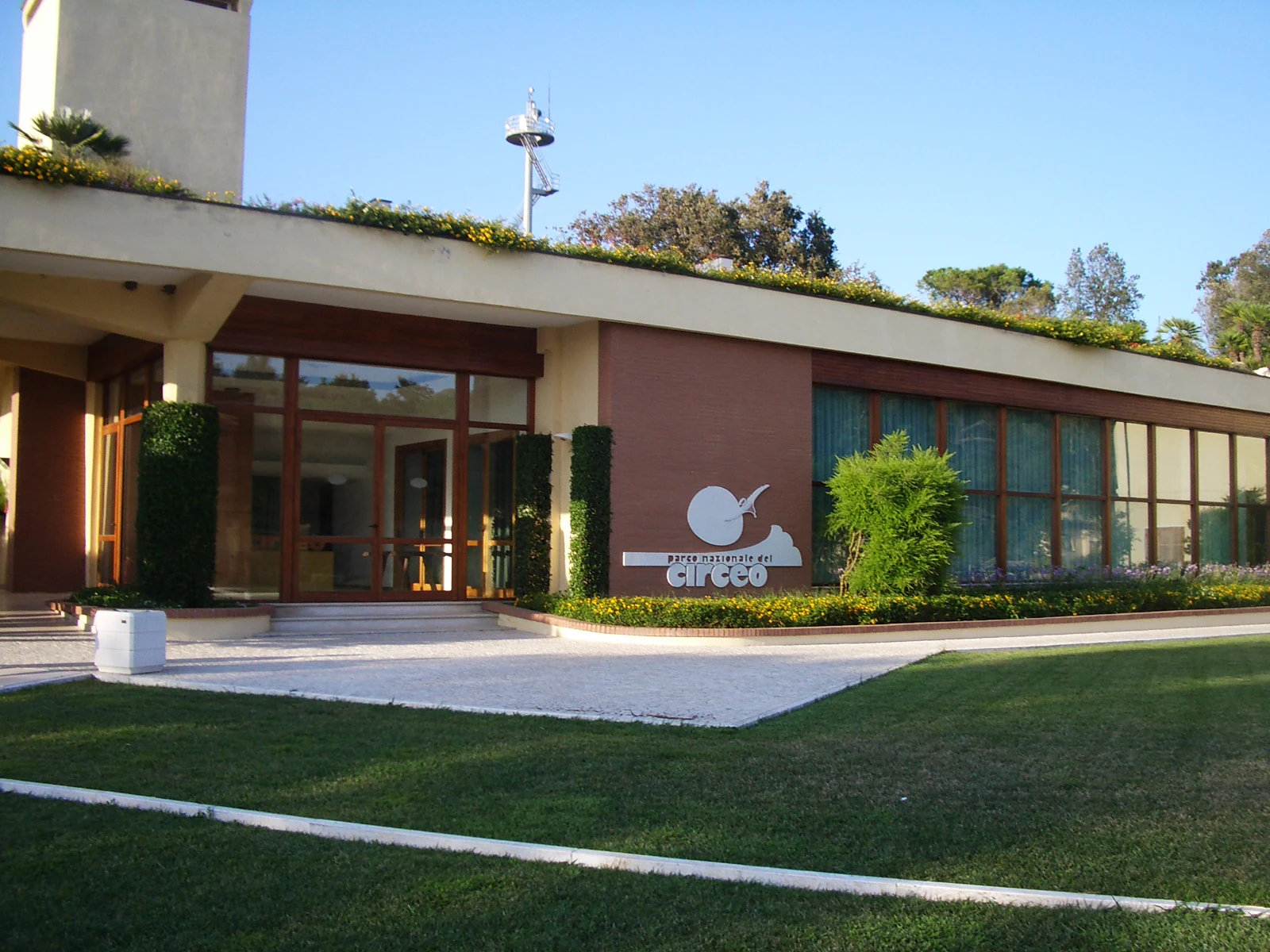 Modern building with glass entrance, beige and brown facade, green roof, and Circeo National Park logo on the wall. The building is surrounded by a well-maintained lawn.