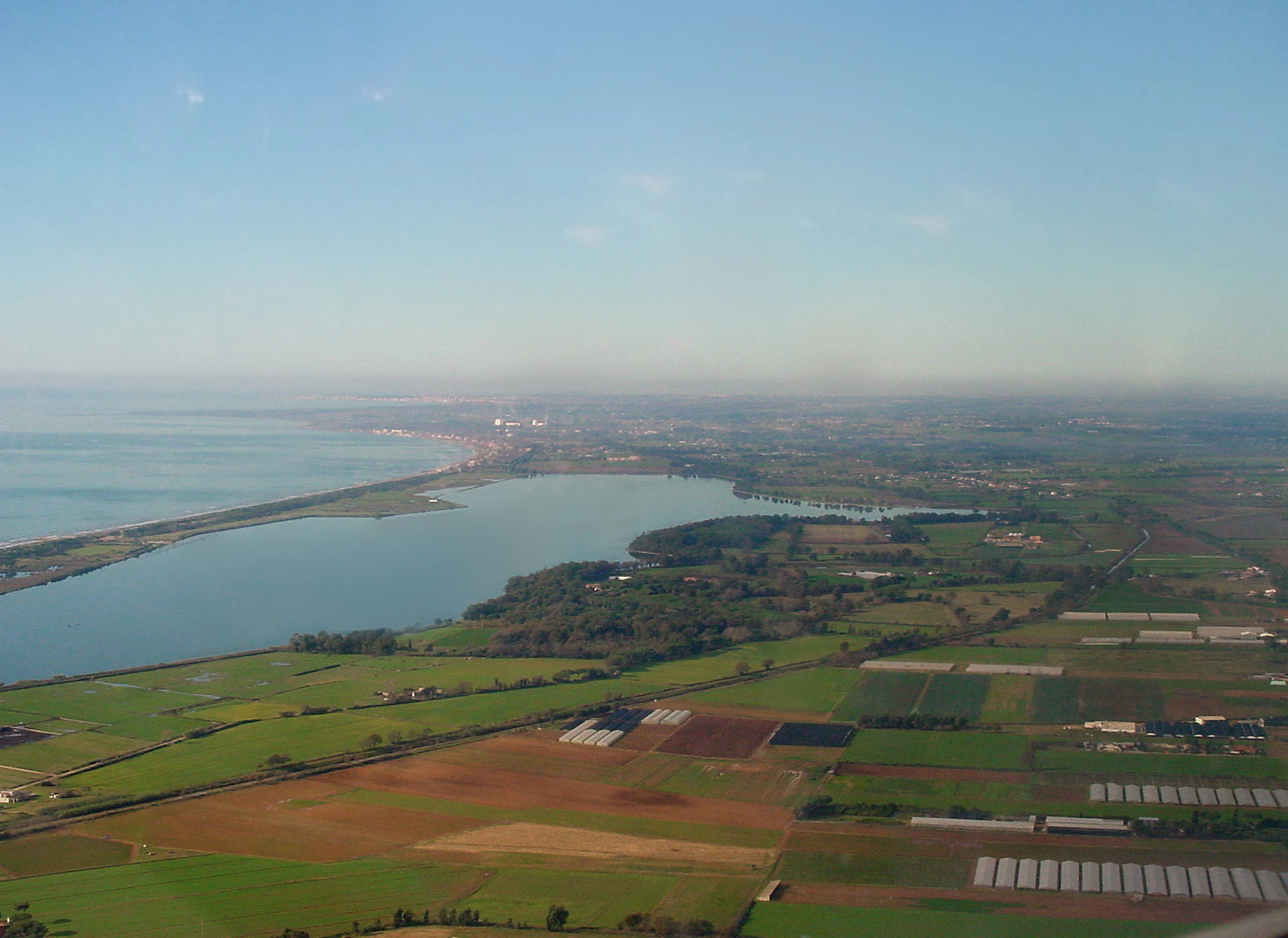 Aerial view of a coastal lake bordered by agricultural fields and sandy shoreline with the sea visible in the background under a clear sky