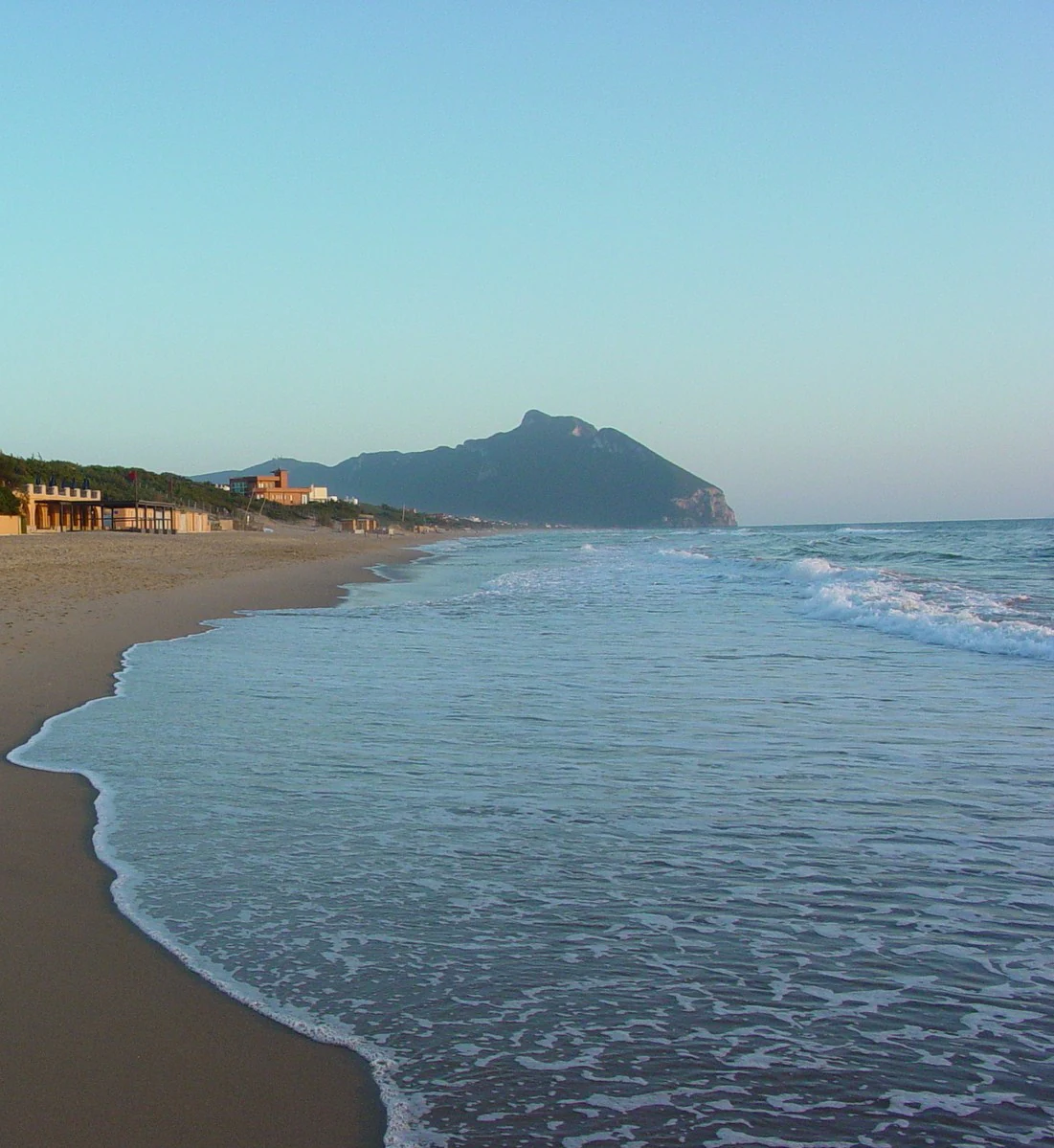 Beach with gentle waves lapping shore, sandy beach, ocean, distant mountain, clear sky, and buildings on the left