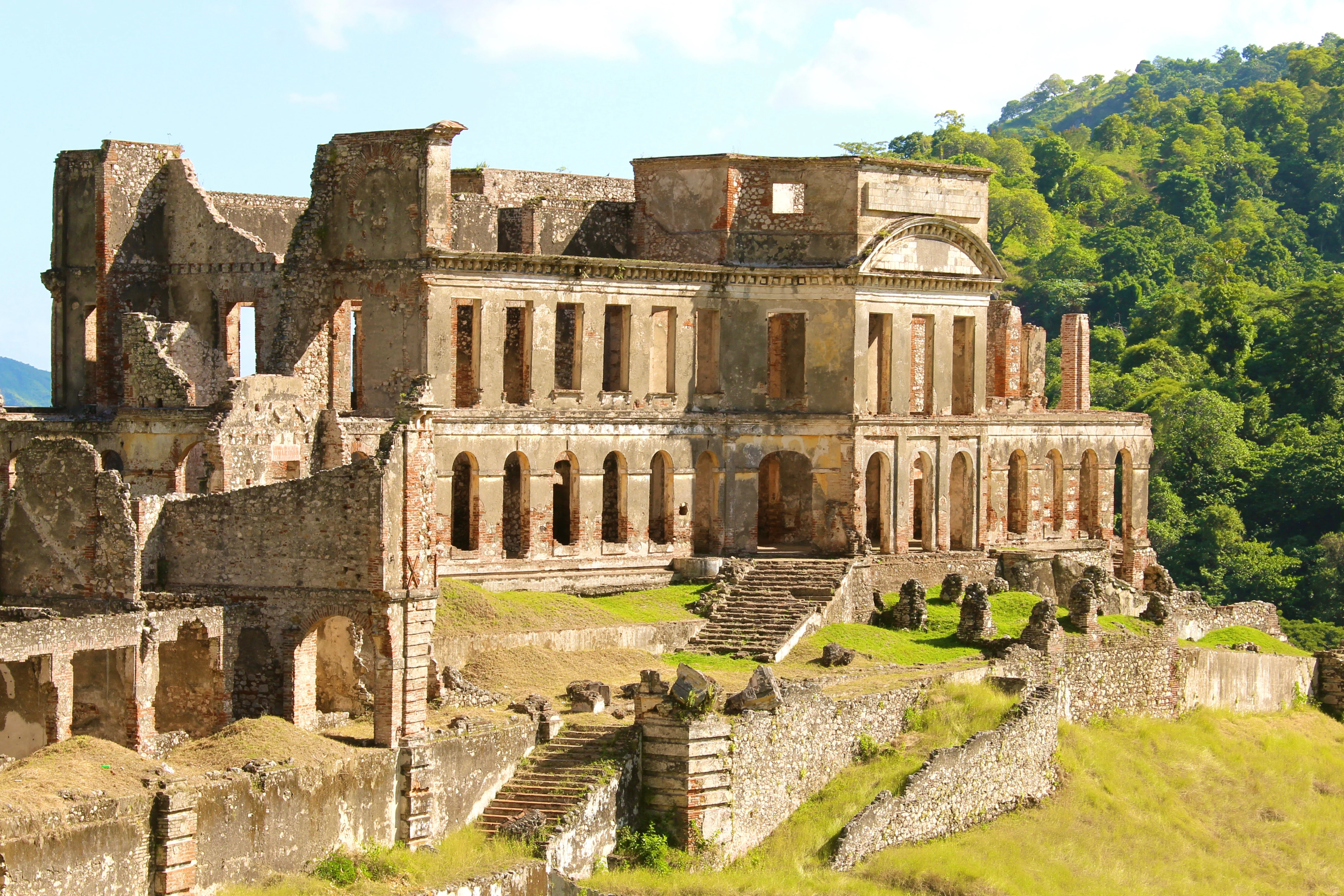 Una vista de gran angular de las ruinas históricas de ladrillo y piedra del Palacio de Sans Souci, que muestra varios niveles de arcos y una gran escalinata central, con un telón de fondo de frondosas colinas boscosas.