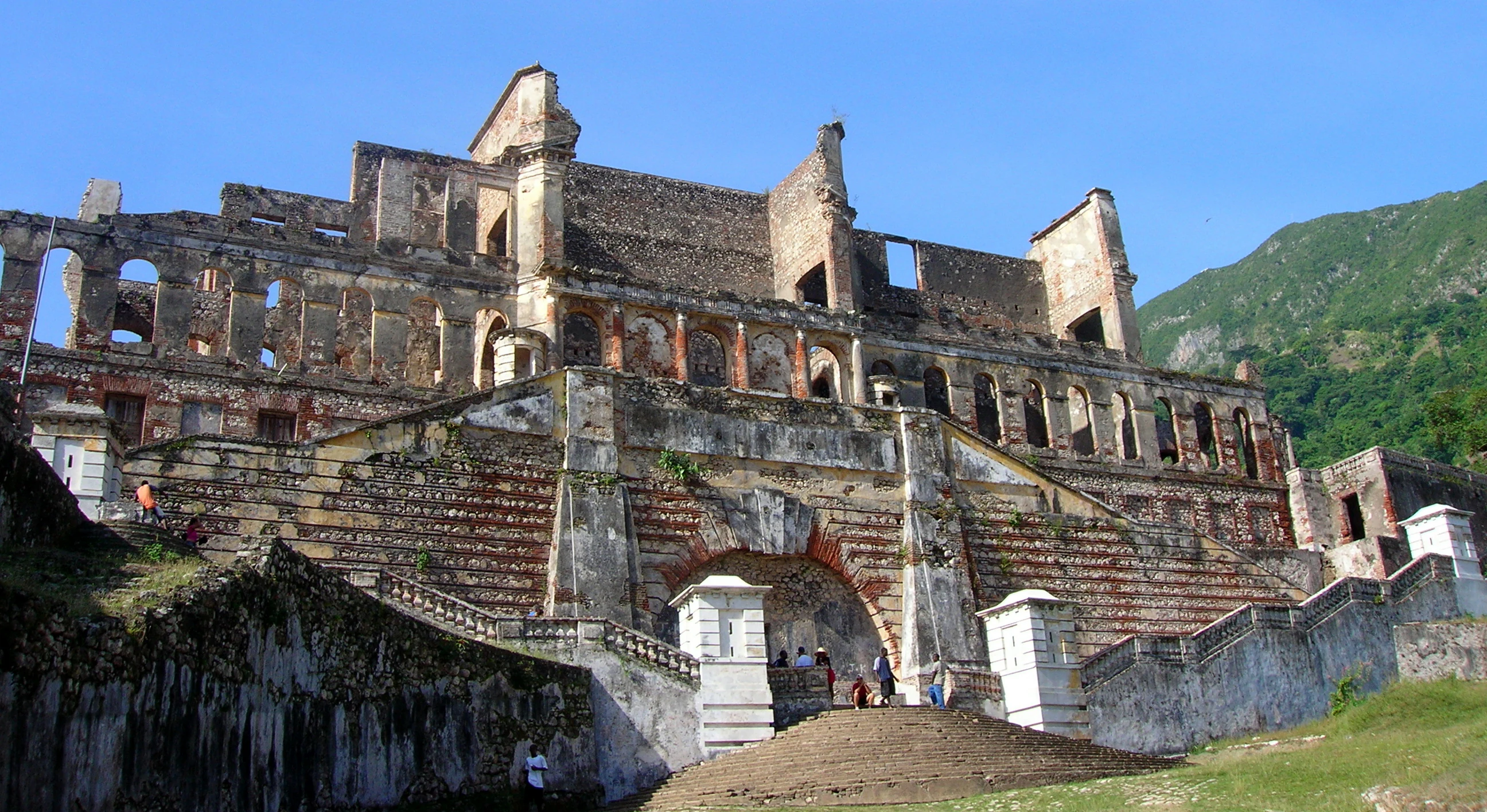 Vista frontal de las ruinas históricas del Palacio de Sans Souci en Haití, mostrando la escalera principal y la fachada de varios niveles contra un cielo azul despejado.