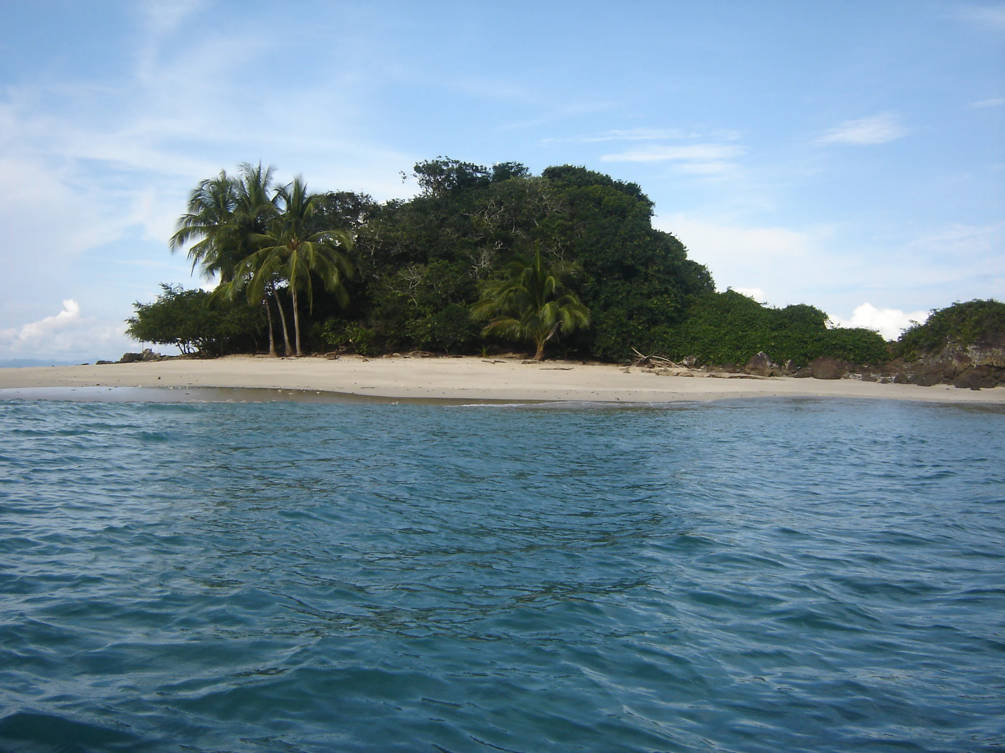 A small, forested tropical island with a sandy beach, surrounded by blue water under a partly cloudy sky.