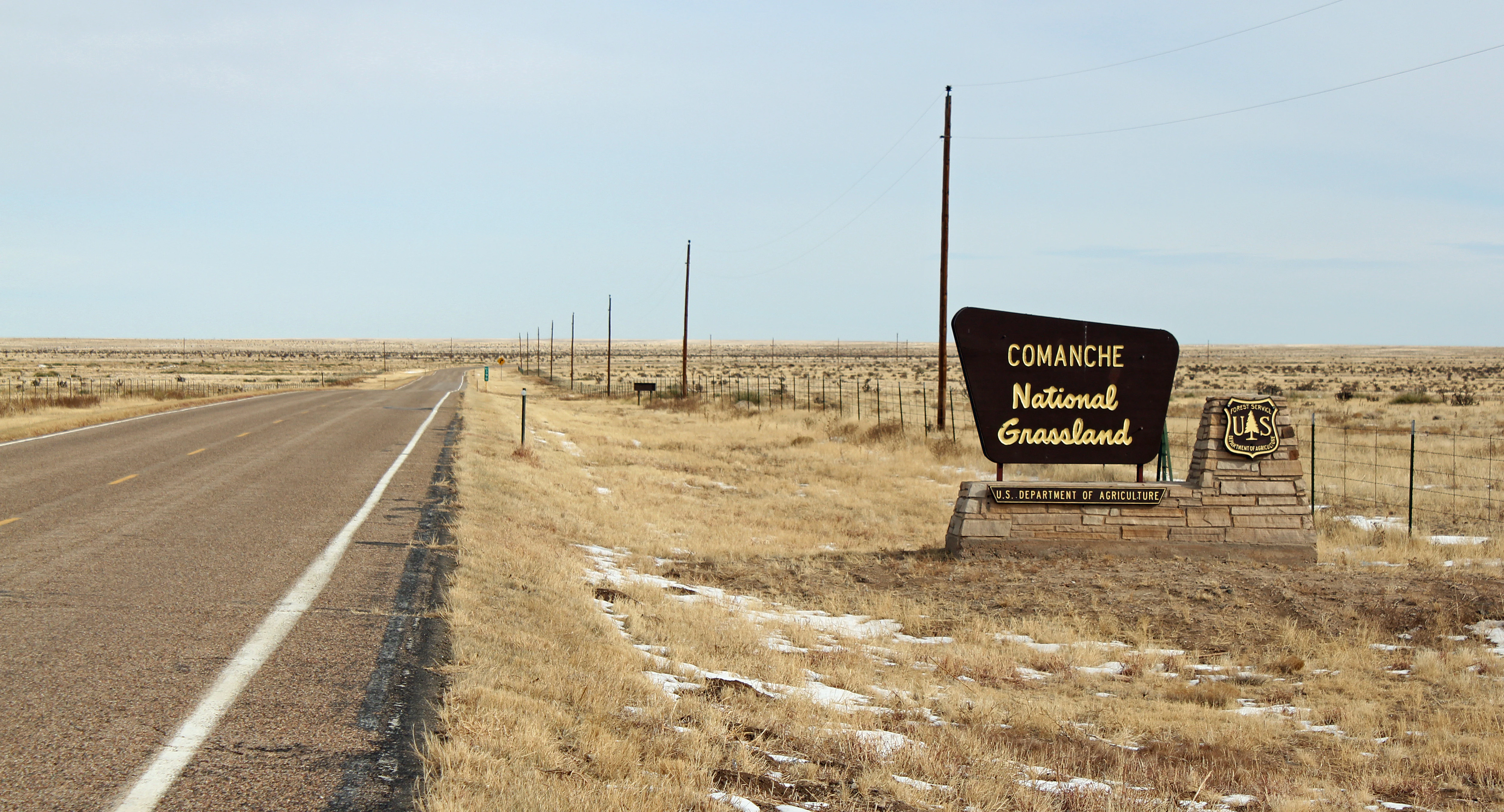A large brown sign reading 'Comanche National Grassland' stands beside a paved road stretching into a flat, grassy landscape with patches of snow.