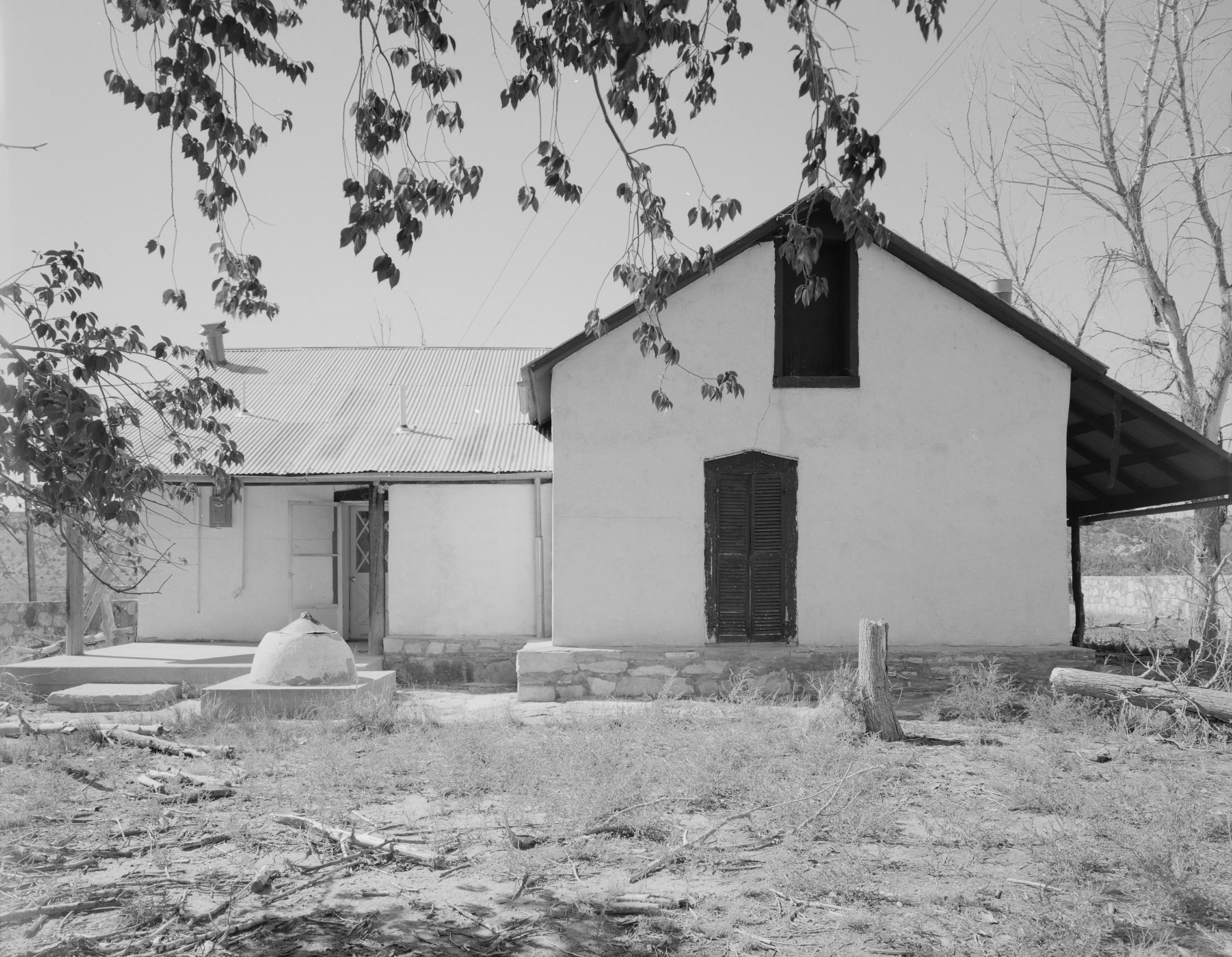 Black and white photograph of the front facade of a historic ranch house with a gabled roof and dark door, surrounded by trees.