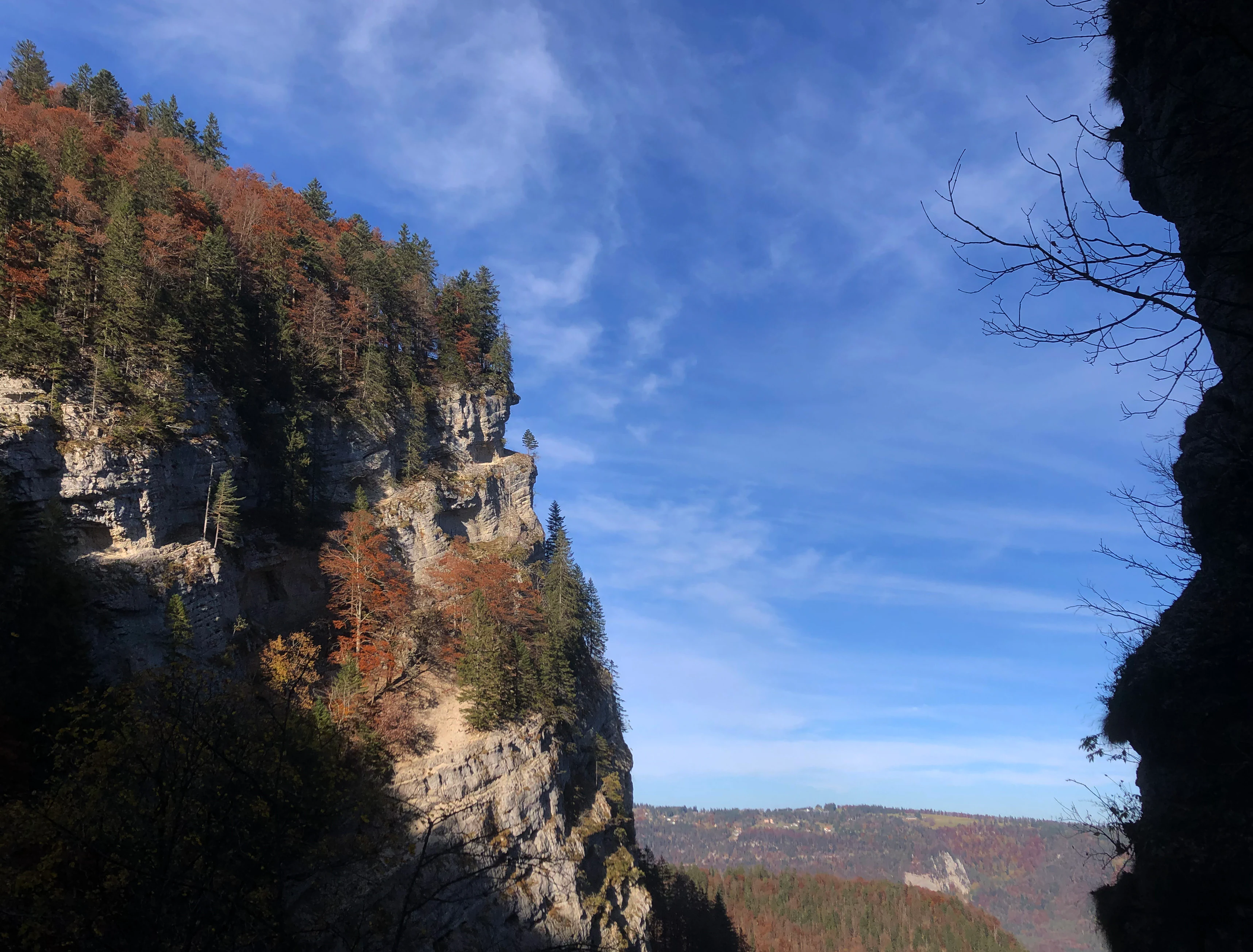 A steep, rocky limestone cliff face with autumn forest vegetation on the left, viewed through a natural gap in the gorge, against a clear blue sky.