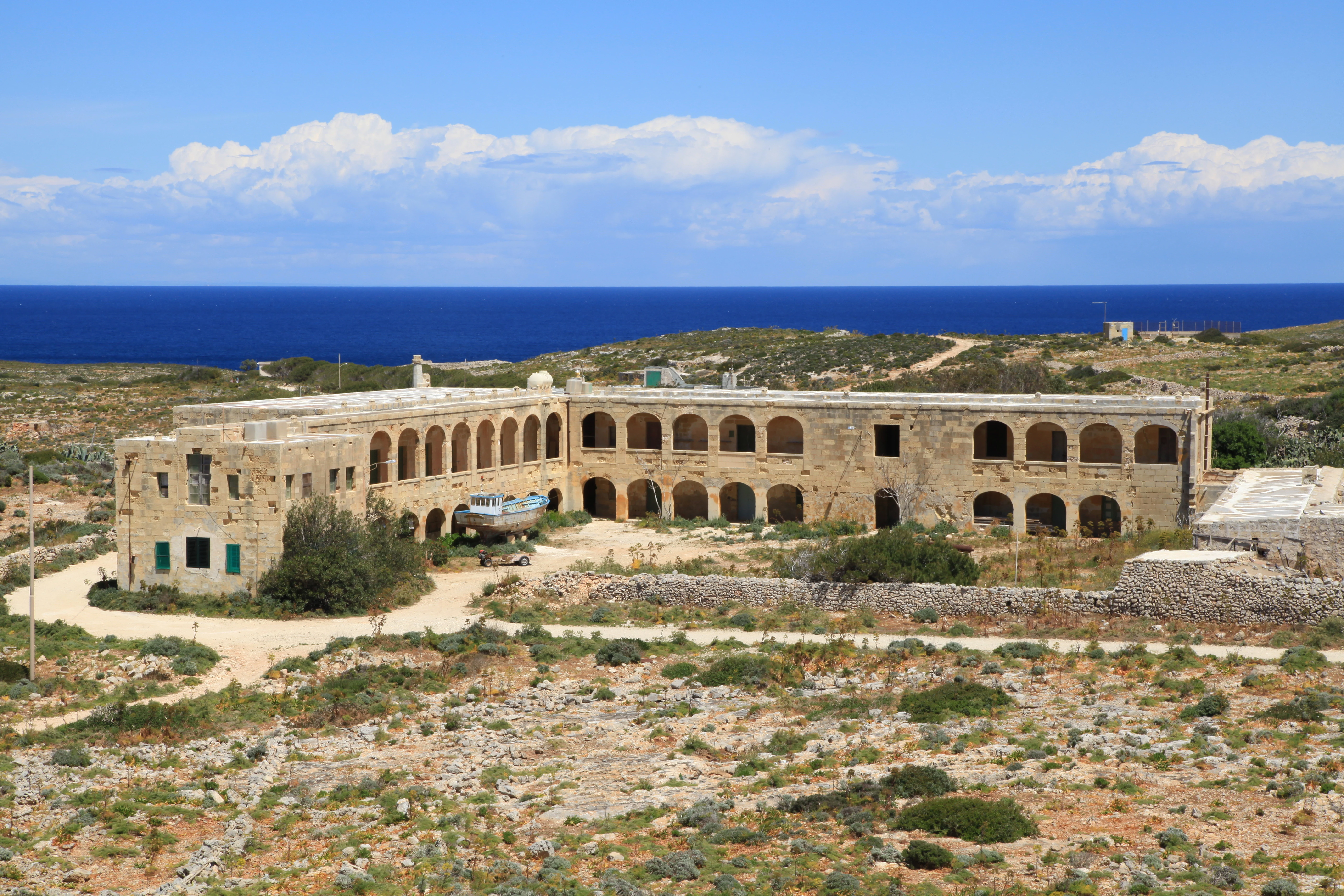 A wide view of a long, two-story stone building with a series of arched windows and doors on a rocky, arid island landscape with the sea in the background.
