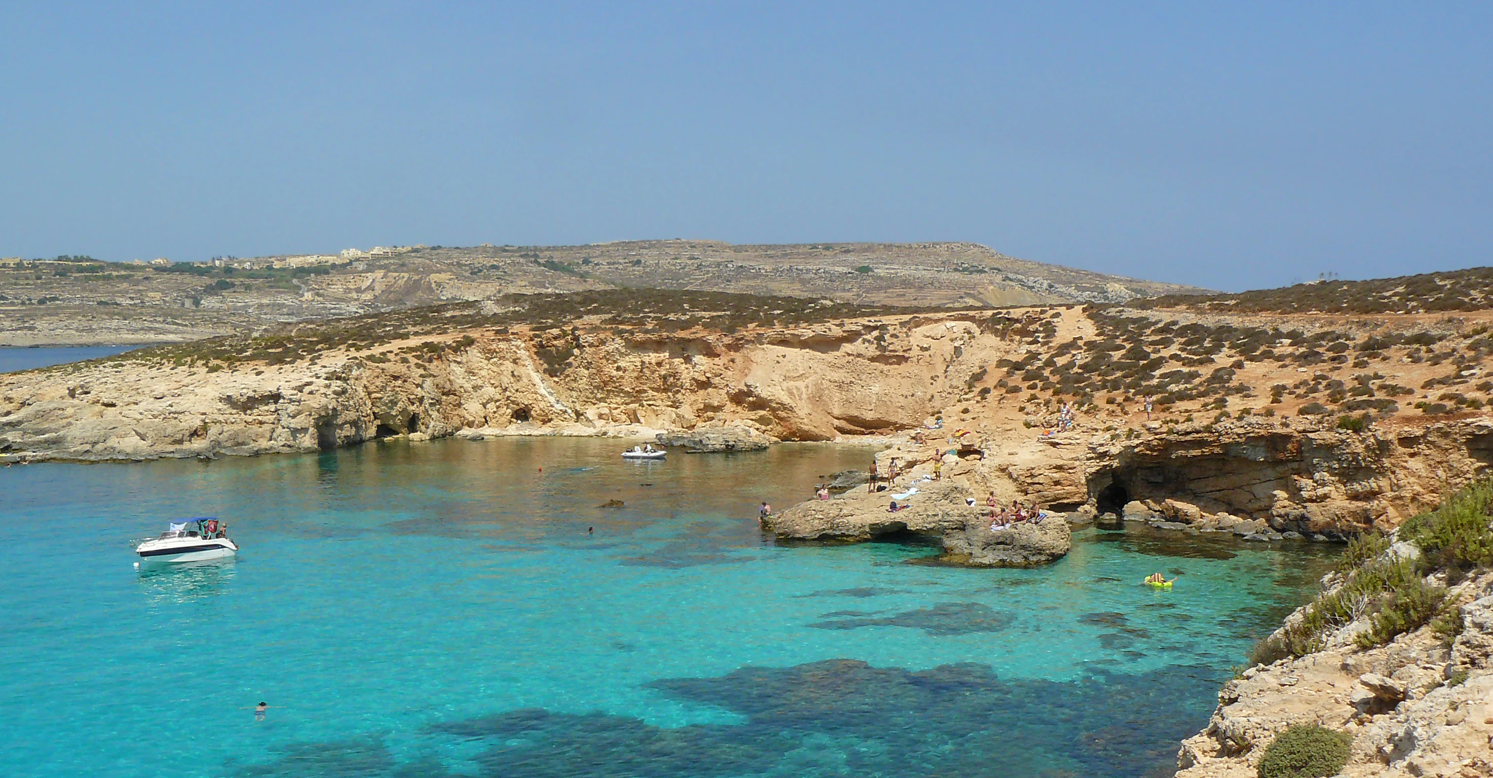 A clear blue lagoon with a small white boat, framed by rocky, arid cliffs and low-lying Mediterranean shrubland on a sunny day.