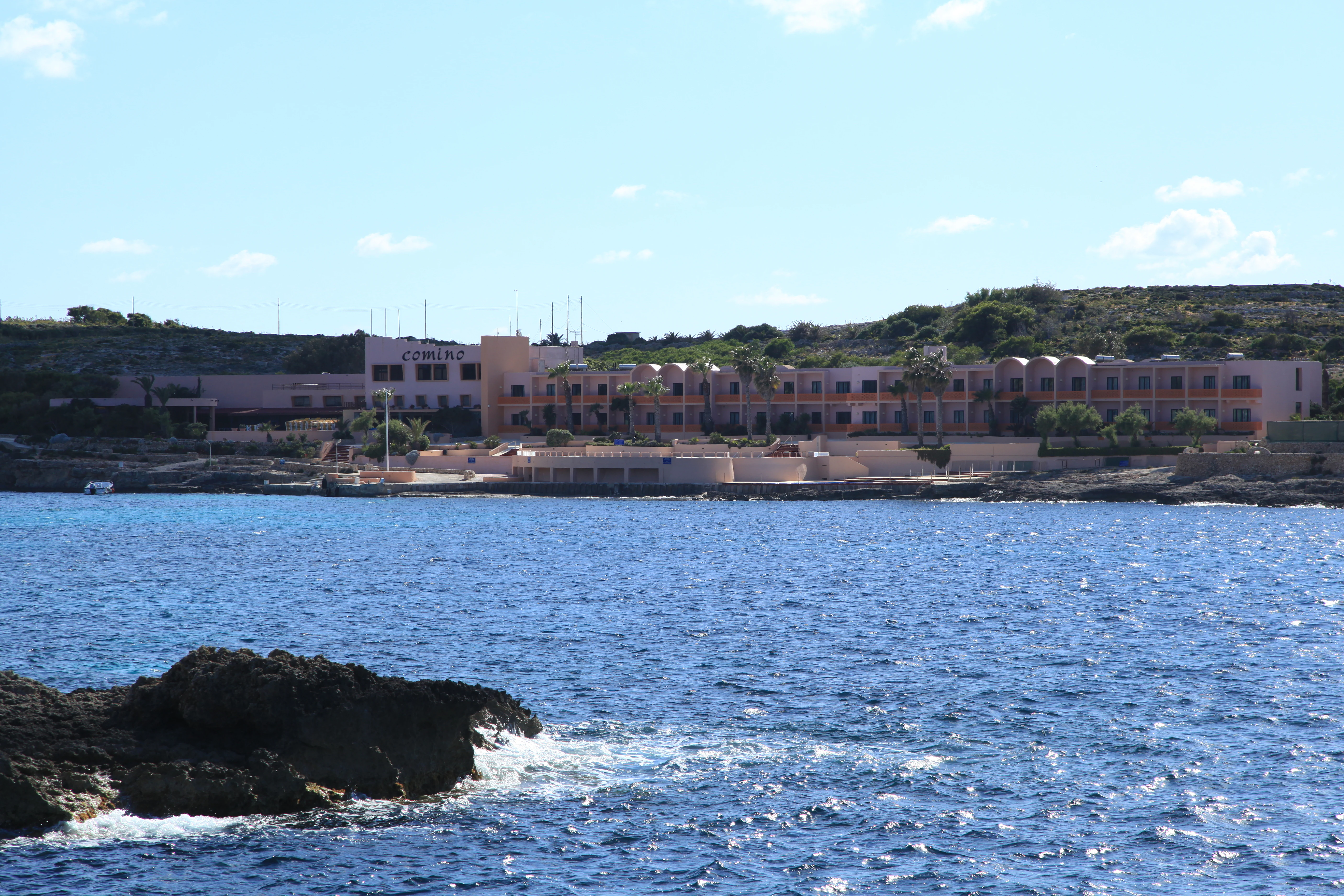 A horizontal view from the water looking toward the Comino Hotel, a low, pinkish-beige building structure set against a rocky hillside on Comino island, Malta.