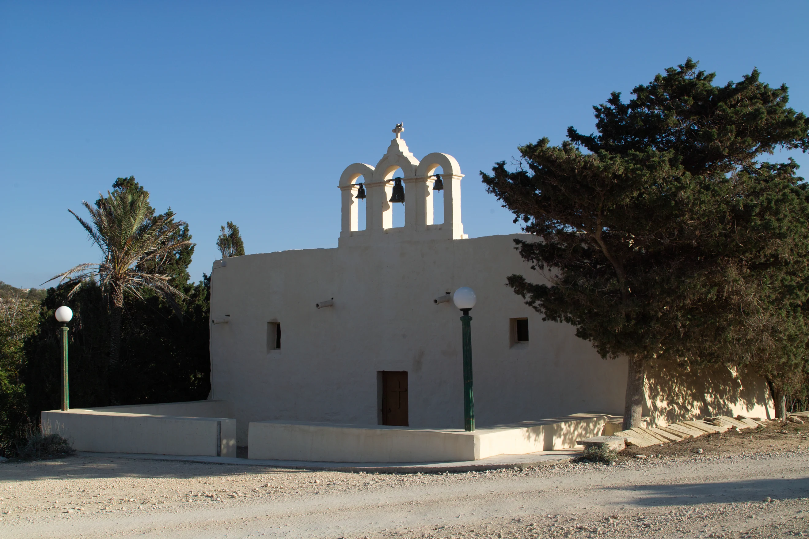 A small light-colored stone chapel with a three-arched bell gable, situated near a palm tree and a larger evergreen tree under a clear sky.