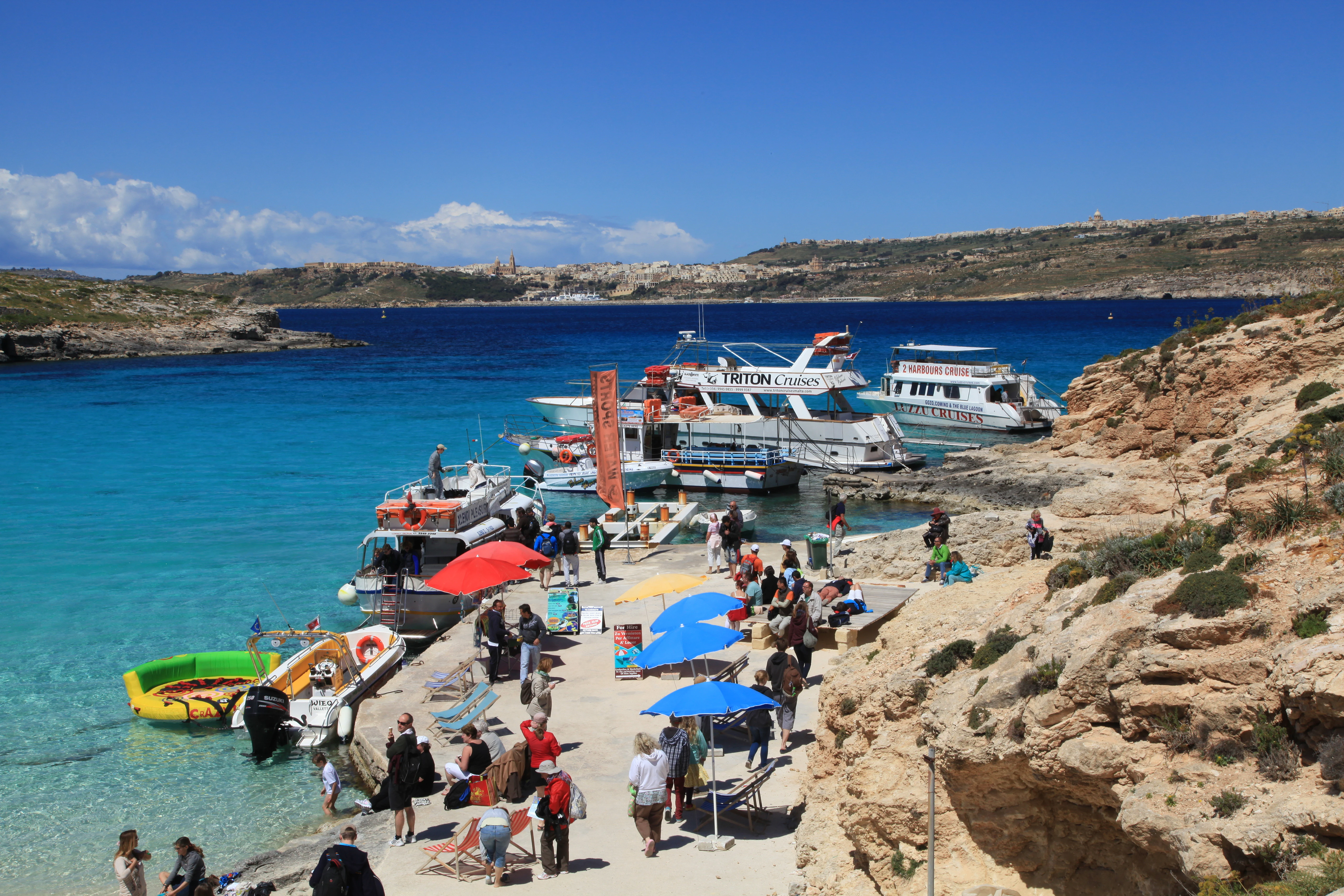 A wide angle view of the Blue Lagoon on Comino island, showing tour boats docked at a small concrete pier, colorful sun umbrellas, and tourists congregating near the rocky shoreline with clear turquoise water.