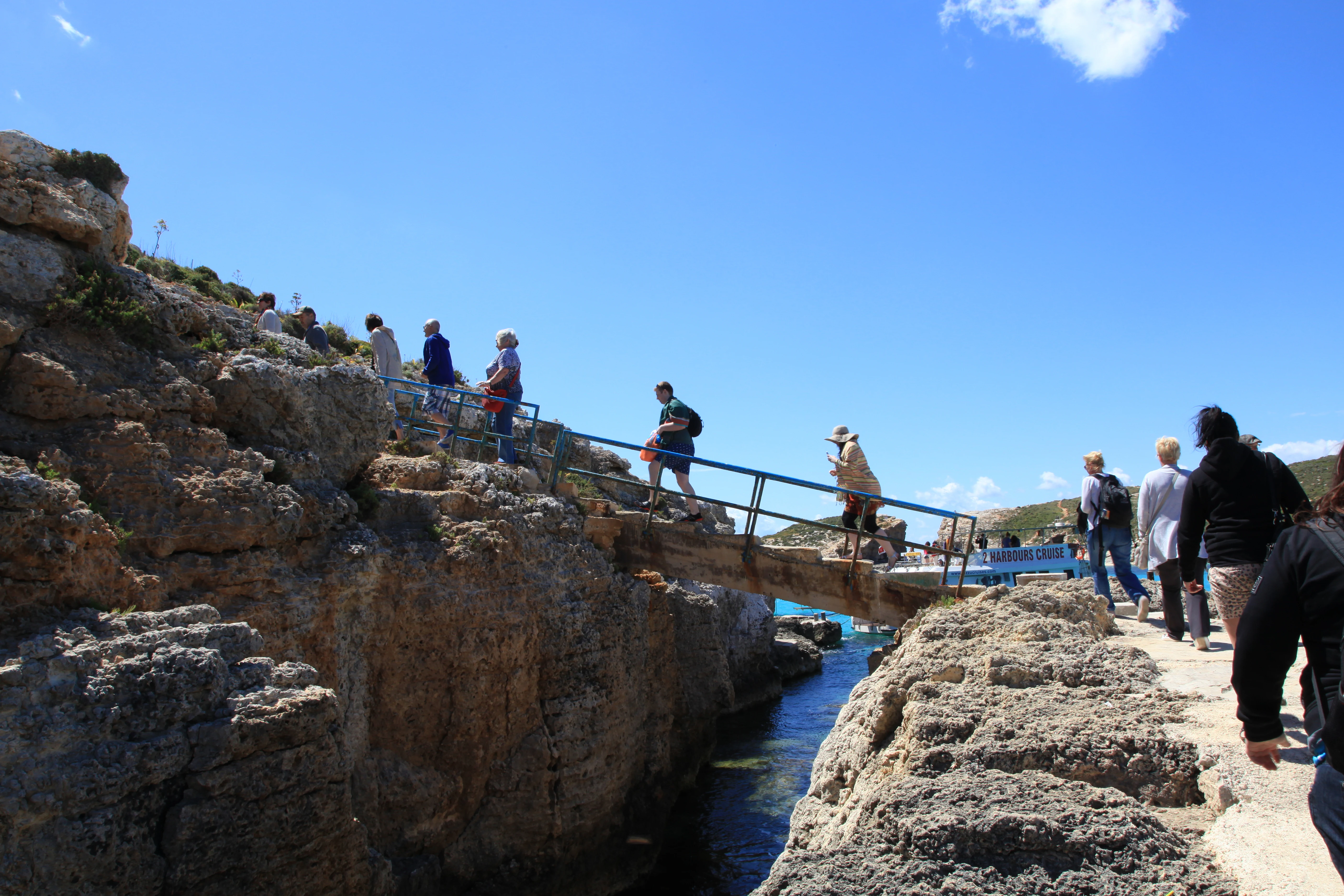 A group of people walking across a small metal footbridge connecting two rocky cliffs over a narrow inlet of water at the Blue Lagoon on Comino.