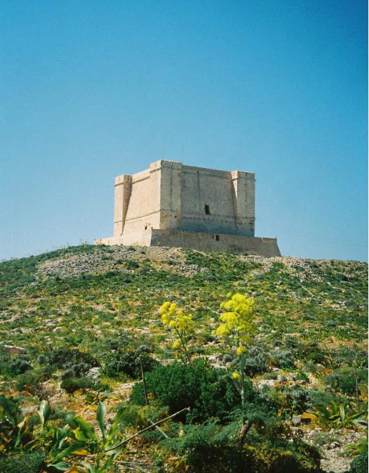 A square stone fortification, St. Mary's Tower, sits atop a grassy, rocky hill under a clear blue sky, with yellow wildflowers in the foreground.