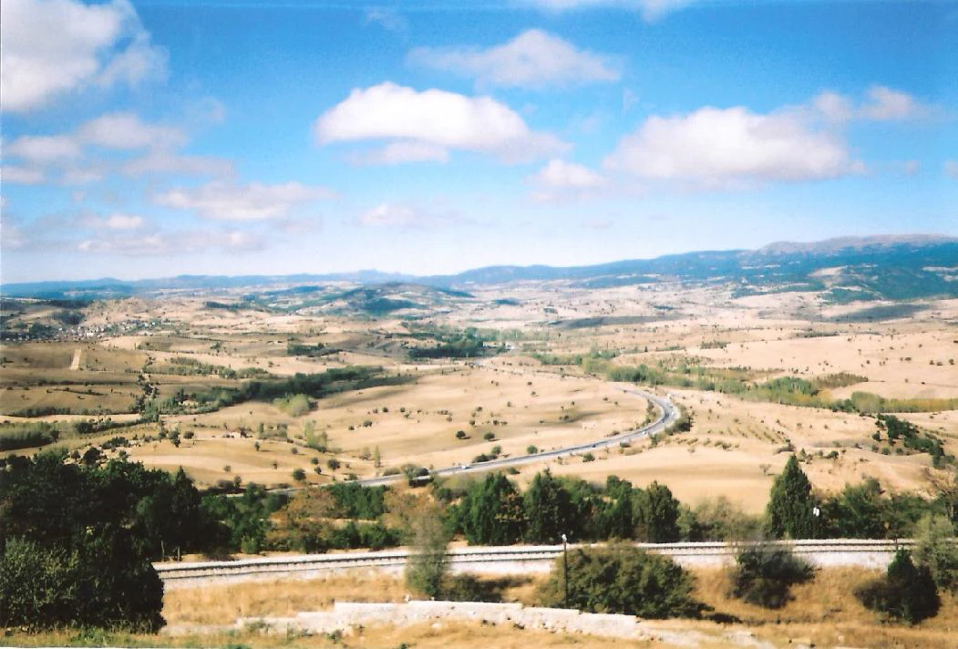 Wide landscape view of dry fields, scattered trees, a curved road, and distant mountains under a blue sky with scattered clouds