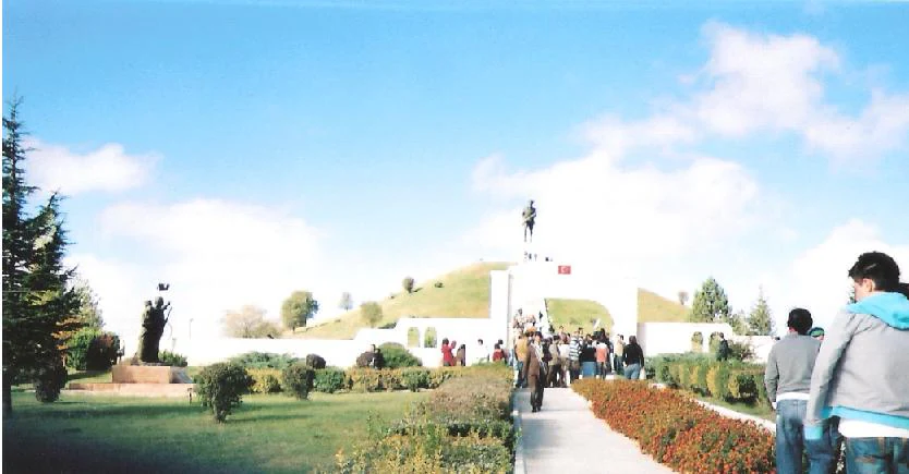 White memorial structure with a statue on a grassy hill, surrounded by pathways, visitors, and landscaped greenery under a blue sky