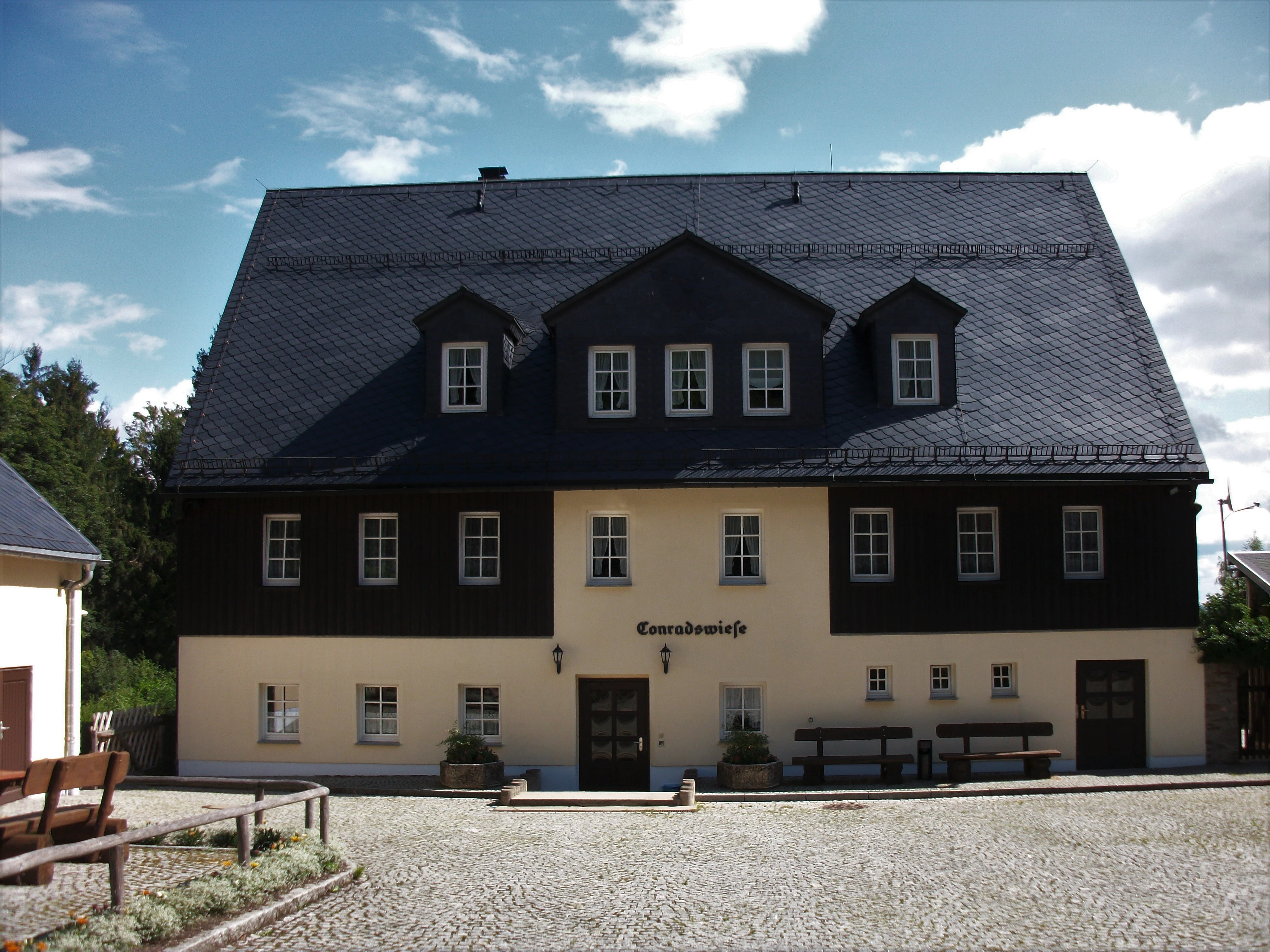 A two-story building with dark wooden siding on the upper floor, cream-colored lower walls, and a large slate-tiled roof, set in a courtyard with a gravel surface.