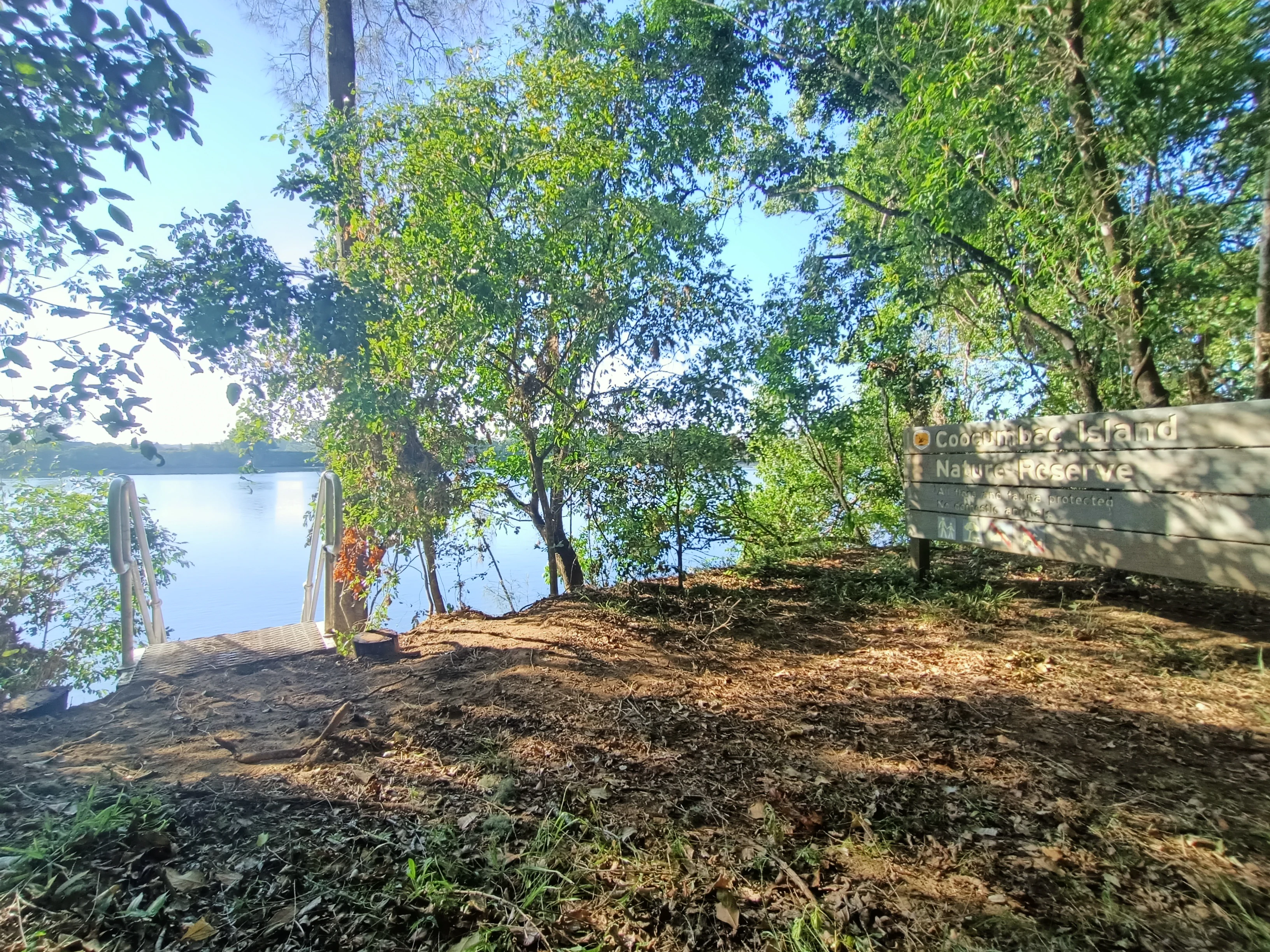 A wooden sign for Coocumbac Island Nature Reserve stands near the edge of the Manning River, with a small staircase leading down to the water surrounded by trees.