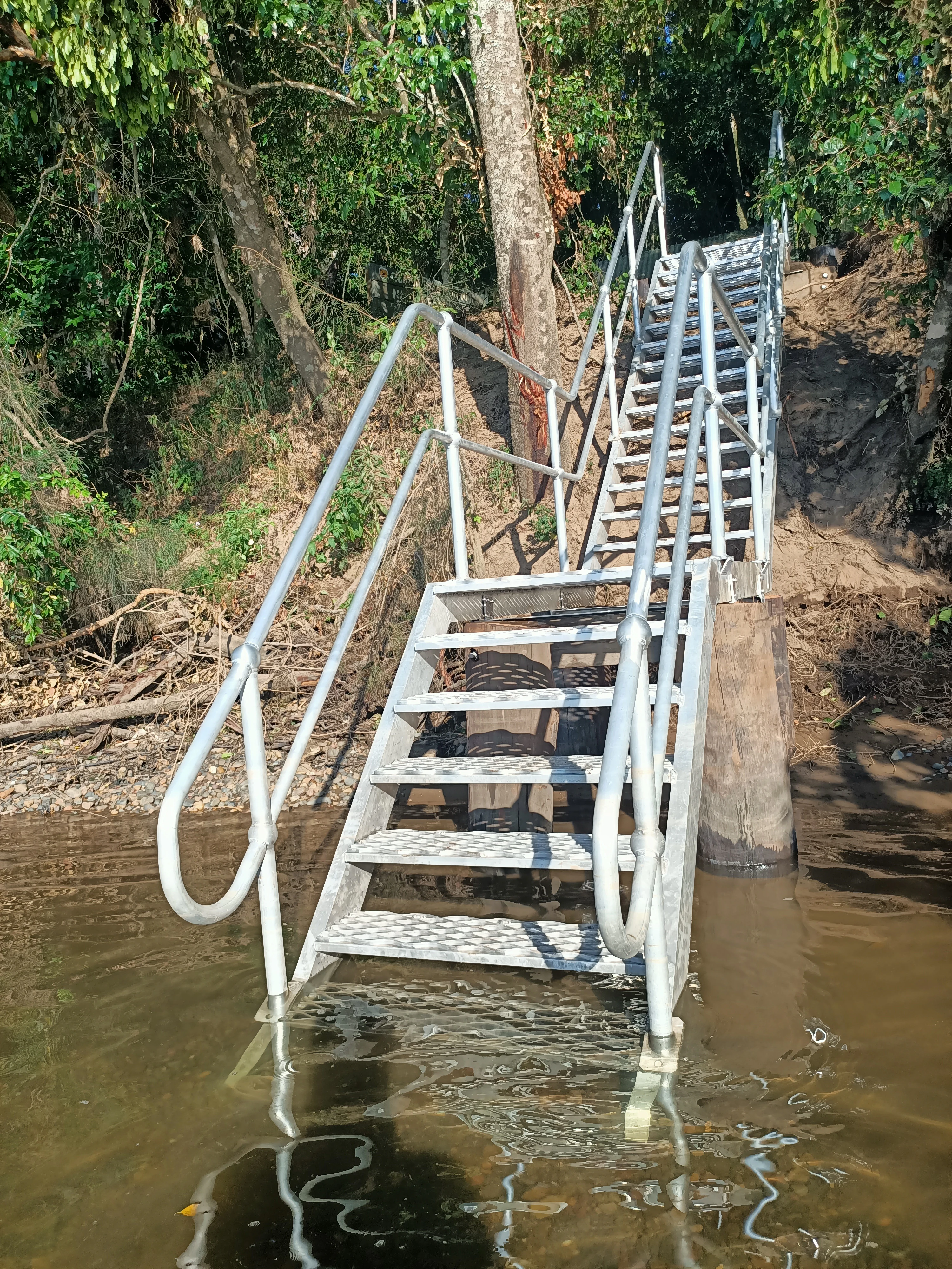 A metal staircase rising out of brown river water, leading up a muddy embankment into a dense forest.