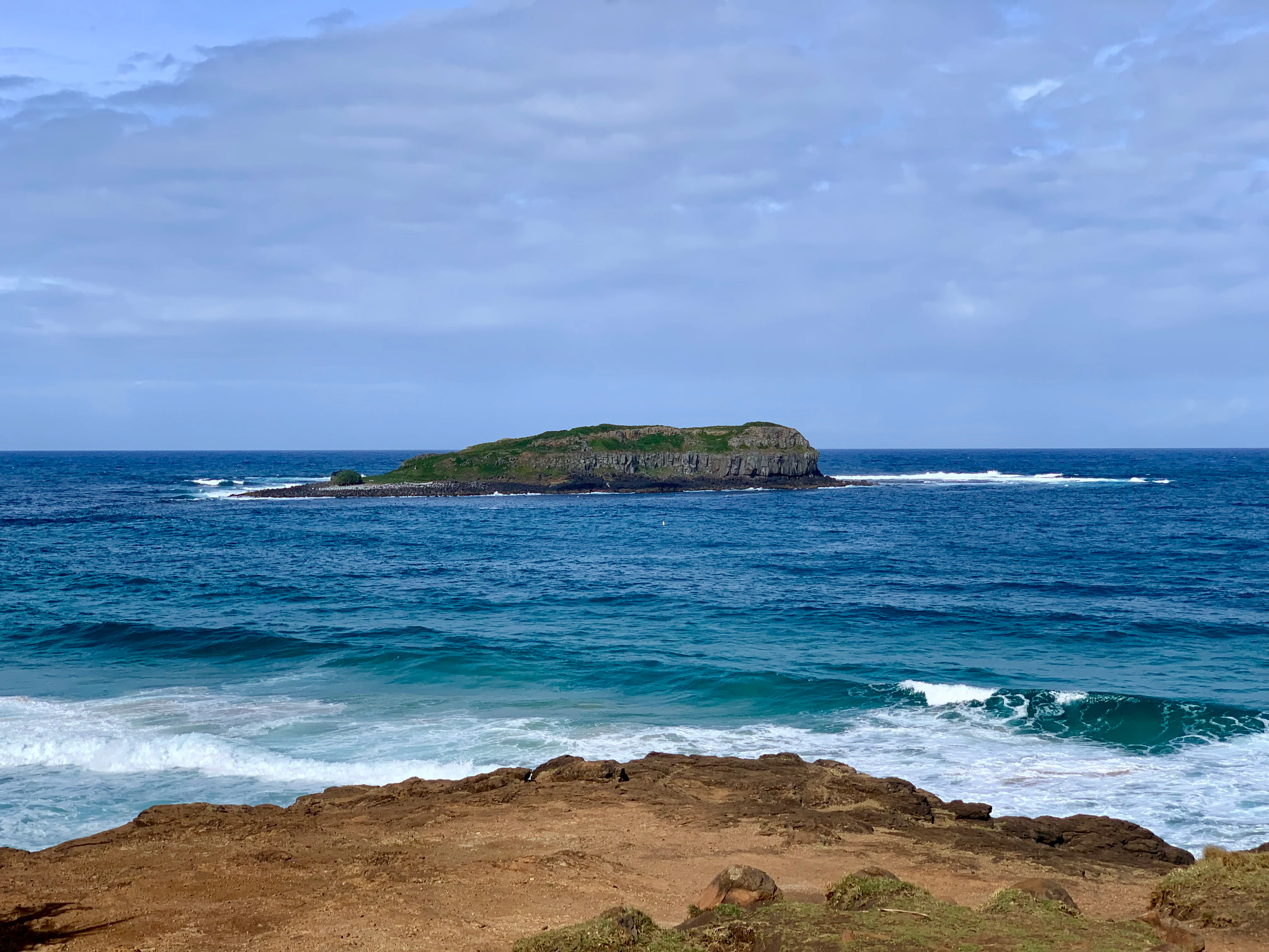 A distant view of a small, green island surrounded by the blue ocean, seen from a rocky coastal vantage point.