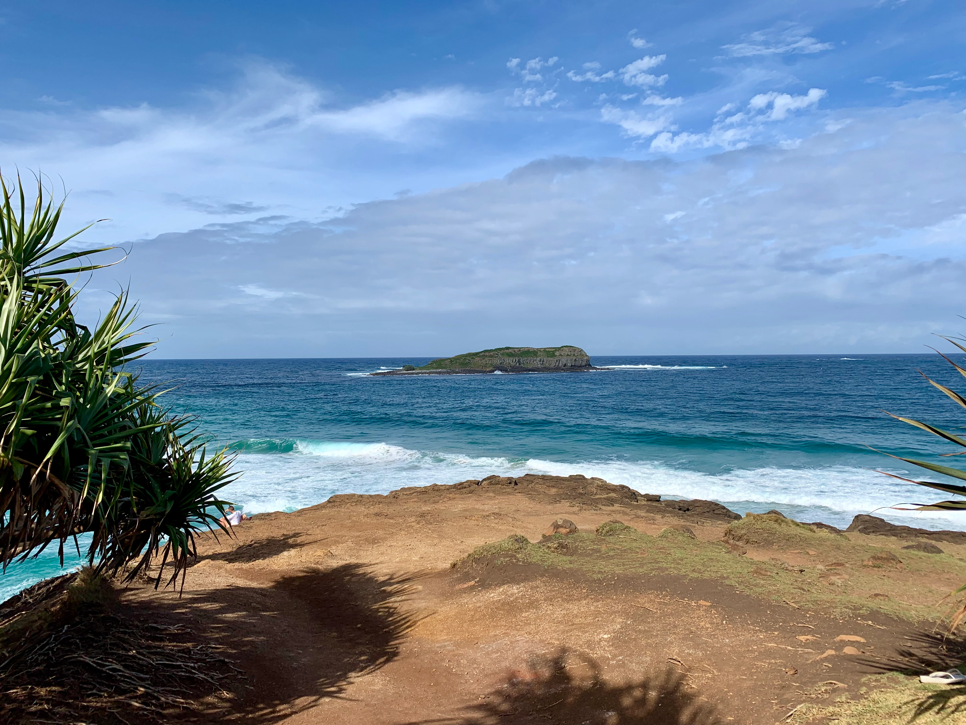 A distant view of Cook Island sitting in the blue ocean, framed by coastal vegetation and a sandy cliffside in the foreground under a partly cloudy sky.