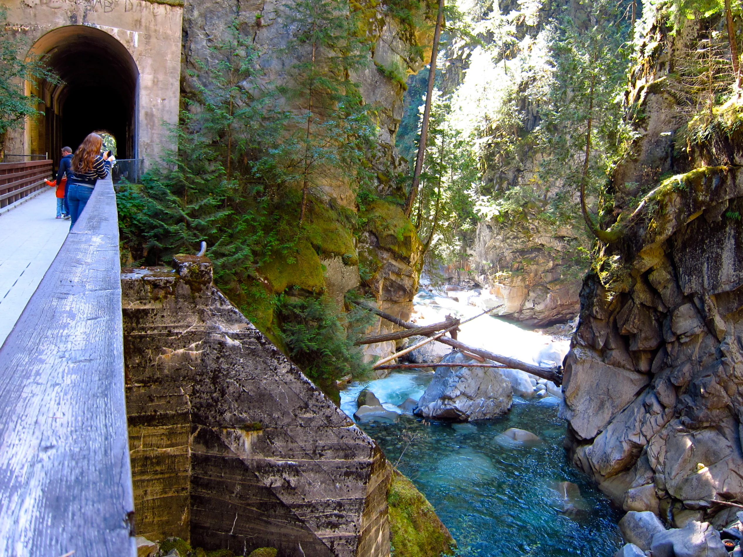 A group of people walking on a wooden walkway alongside a stone tunnel entrance at the edge of a deep, rocky canyon with a turquoise river flowing below.