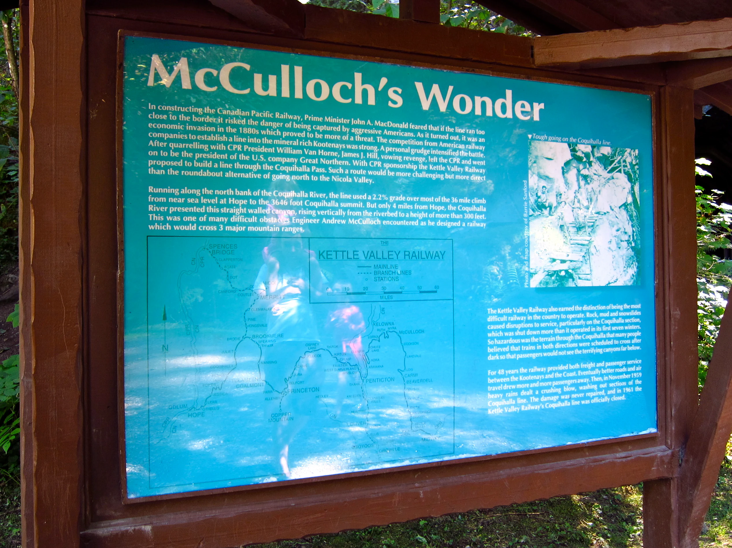 A rectangular information sign made of blue metal with white text and a map, mounted on a wooden frame, explaining the history of the Kettle Valley Railway.