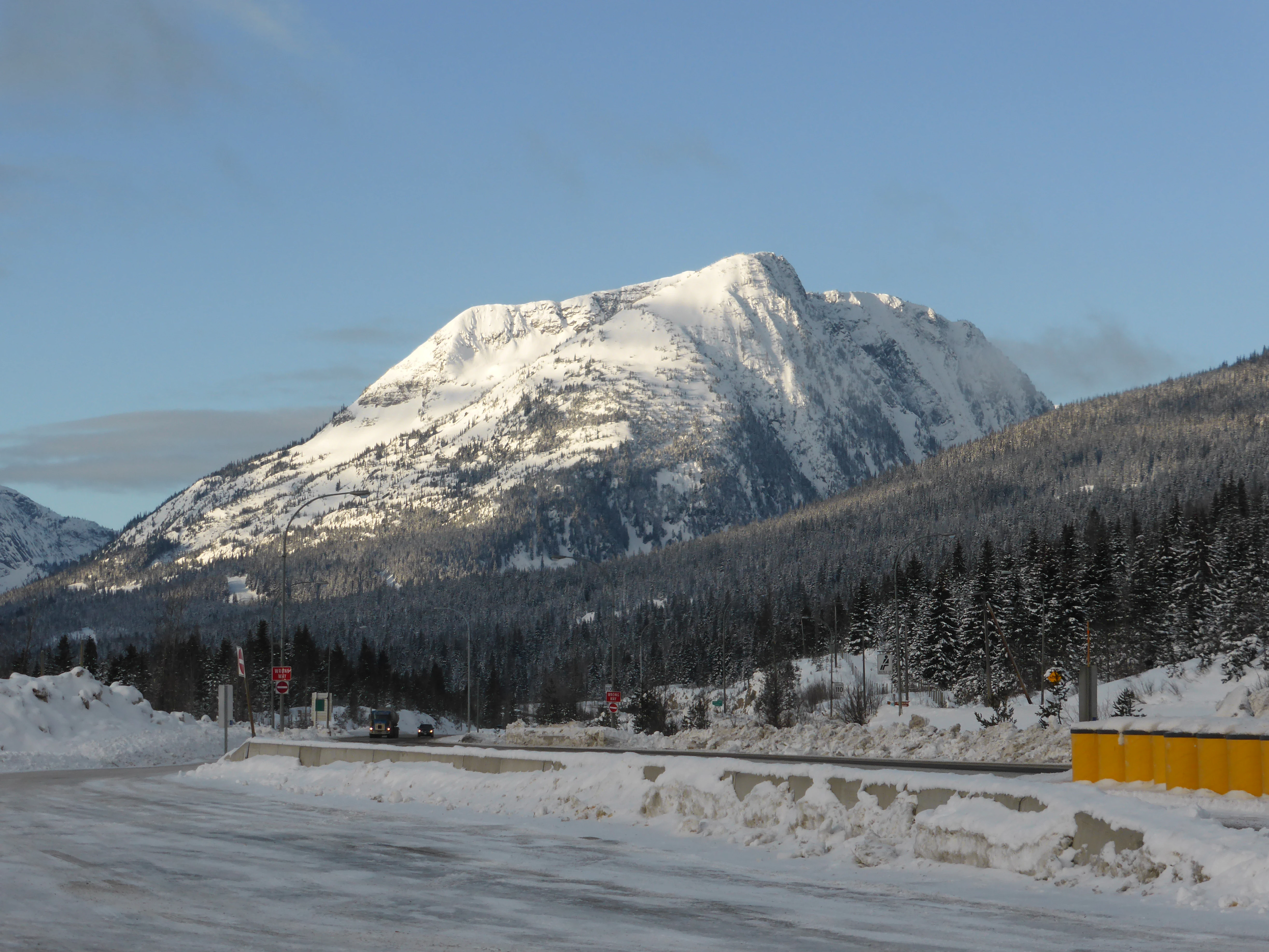 Un pico montañoso cubierto de nieve se eleva sobre un bosque de árboles perennes, con una carretera nevada y una barrera amarilla en primer plano.