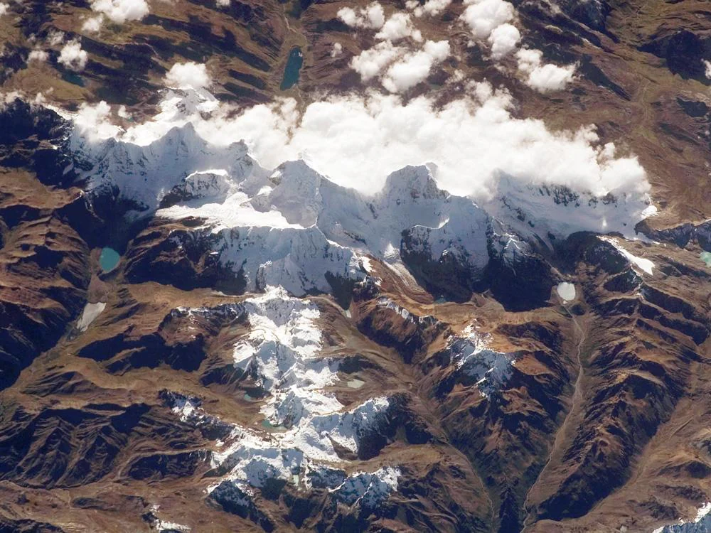 An overhead satellite image of the Cordillera Huayhuash showing mountain peaks, glaciers, alpine lakes, and cloud cover over a brown, rugged landscape.