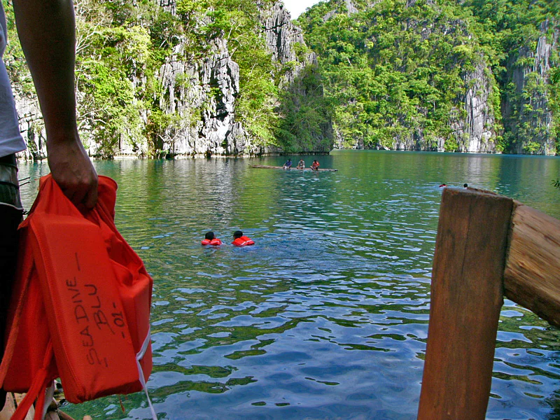 A first person perspective of someone holding a bright orange life vest, looking out over a calm, turquoise lake surrounded by steep, jungle-covered limestone cliffs.
