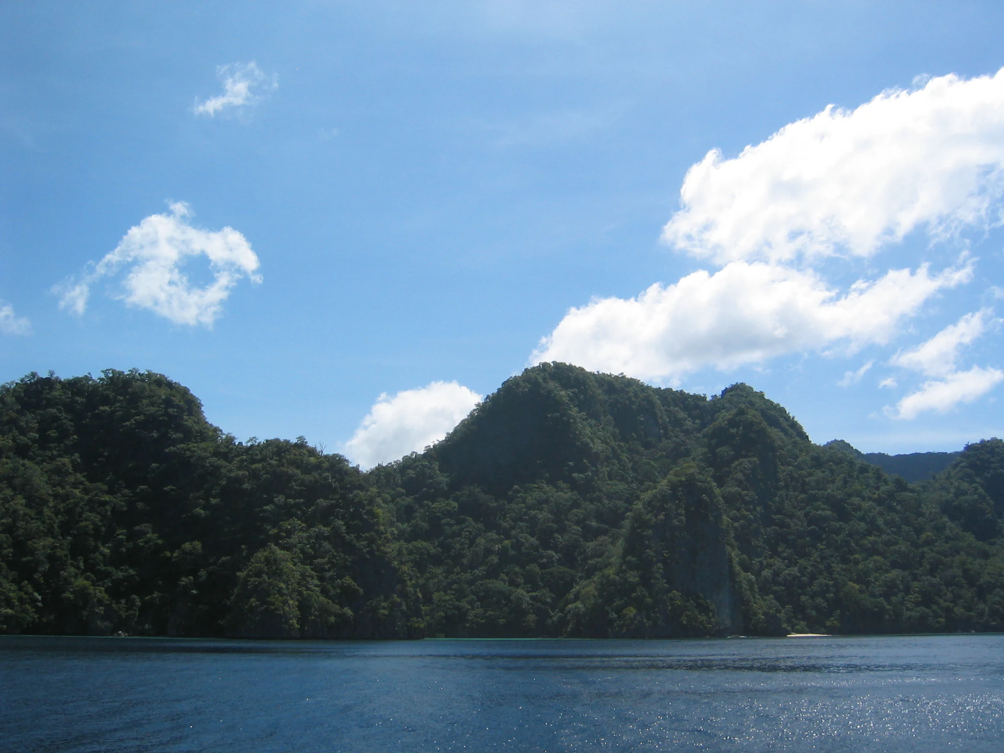 A wide view of tropical karst limestone islands covered in dense green forest rising directly out of dark blue water under a bright blue sky with scattered clouds.