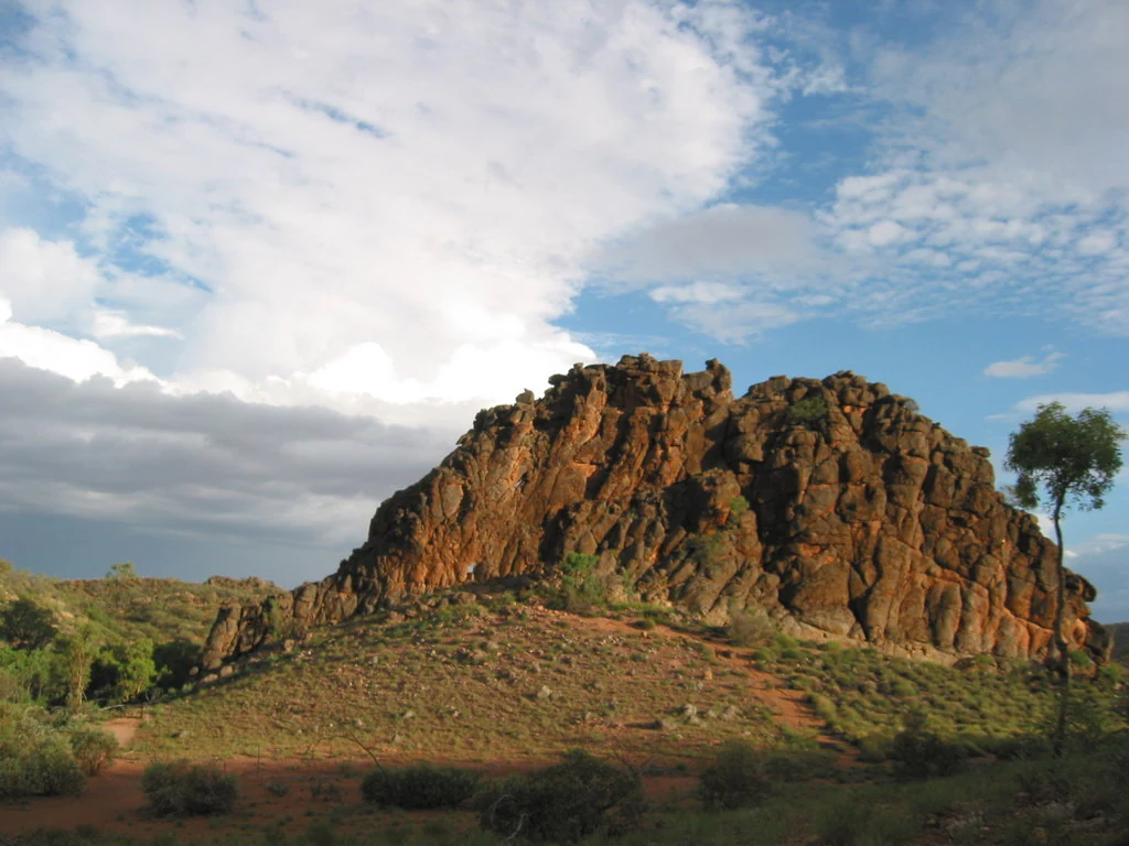 A large, jagged outcrop of dark orange and grey dolomite rock sits prominently in a desert landscape under a blue sky with scattered clouds.