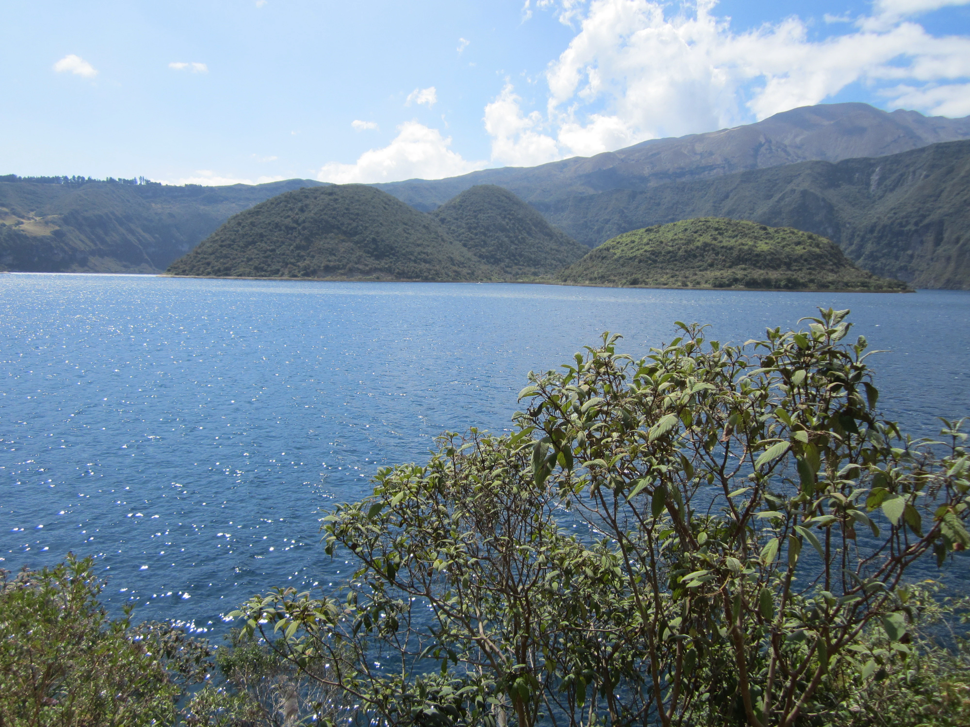 A wide view of a deep blue crater lake with grassy islands in the center, surrounded by mountainous terrain under a partly cloudy sky.