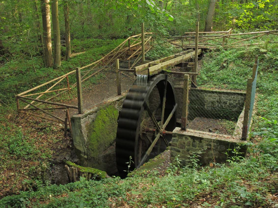 An overshot water wheel in a wooded area, with wooden railings surrounding the structure and forest foliage in the background.