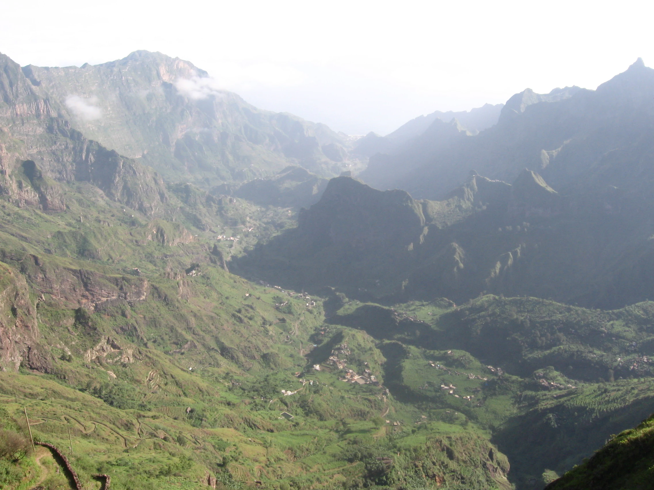 Vast green valley with rugged mountain slopes, scattered settlements, and winding paths viewed from a high vantage point