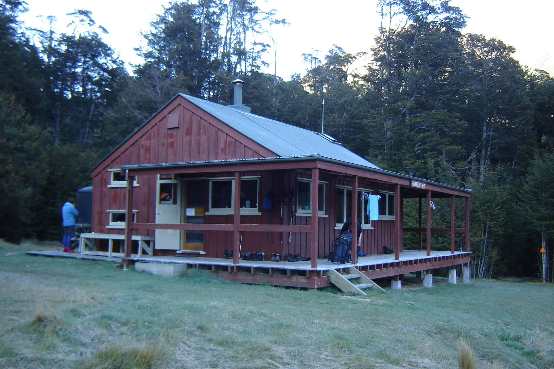 A red wooden backcountry hut with a covered porch sits on a grassy clearing at the edge of a forest.