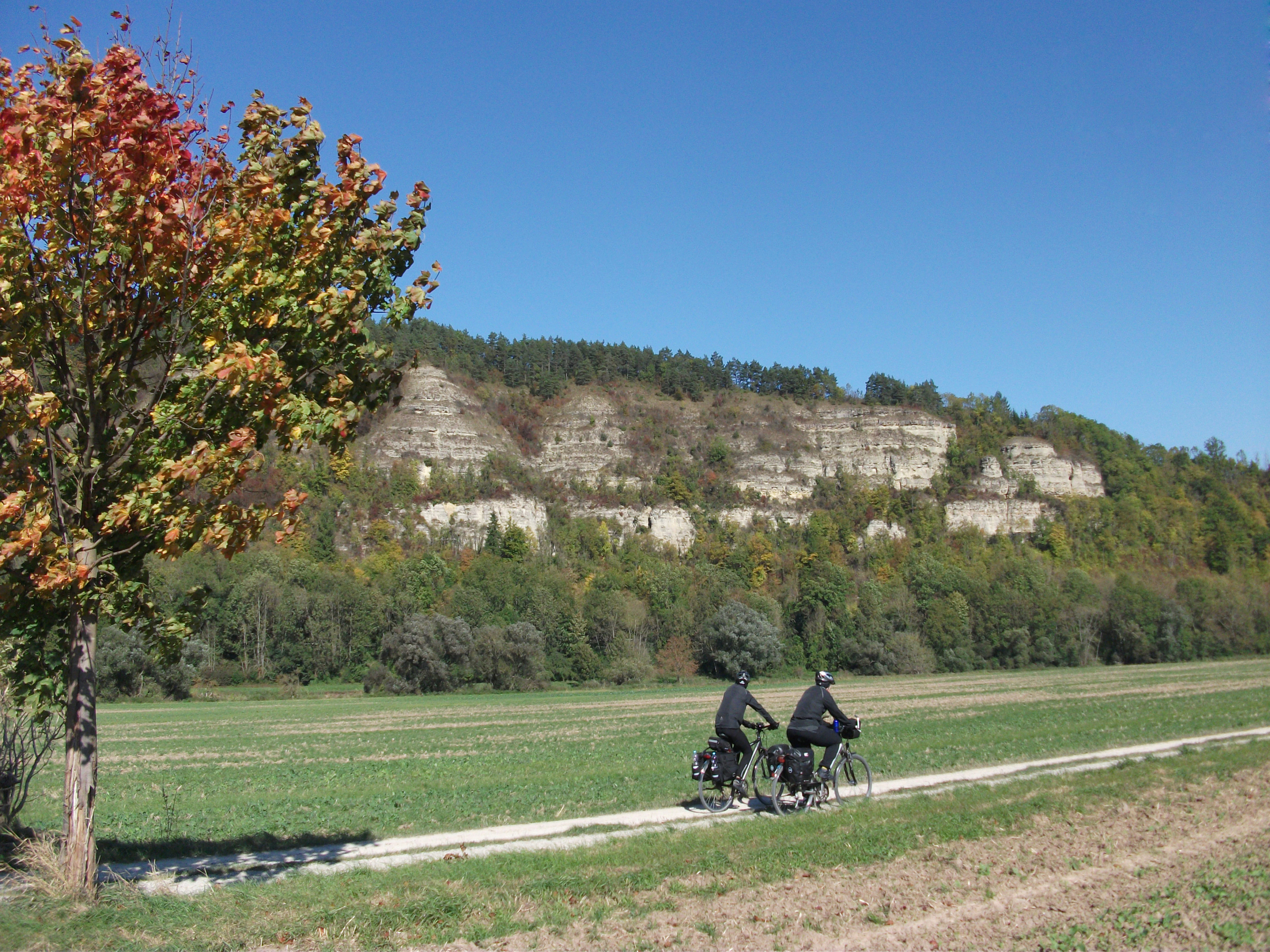 Two cyclists riding on a paved path through a flat field with a steep, forested limestone cliff face in the background under a clear blue sky.