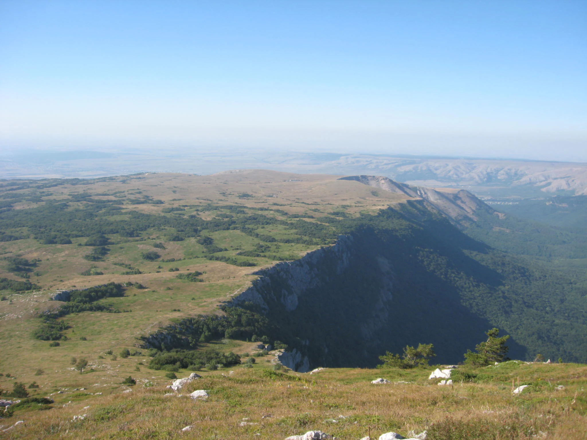 Wide landscape view of a mountainous plateau with grassy meadows, rocky cliffs, and distant valleys under a clear blue sky