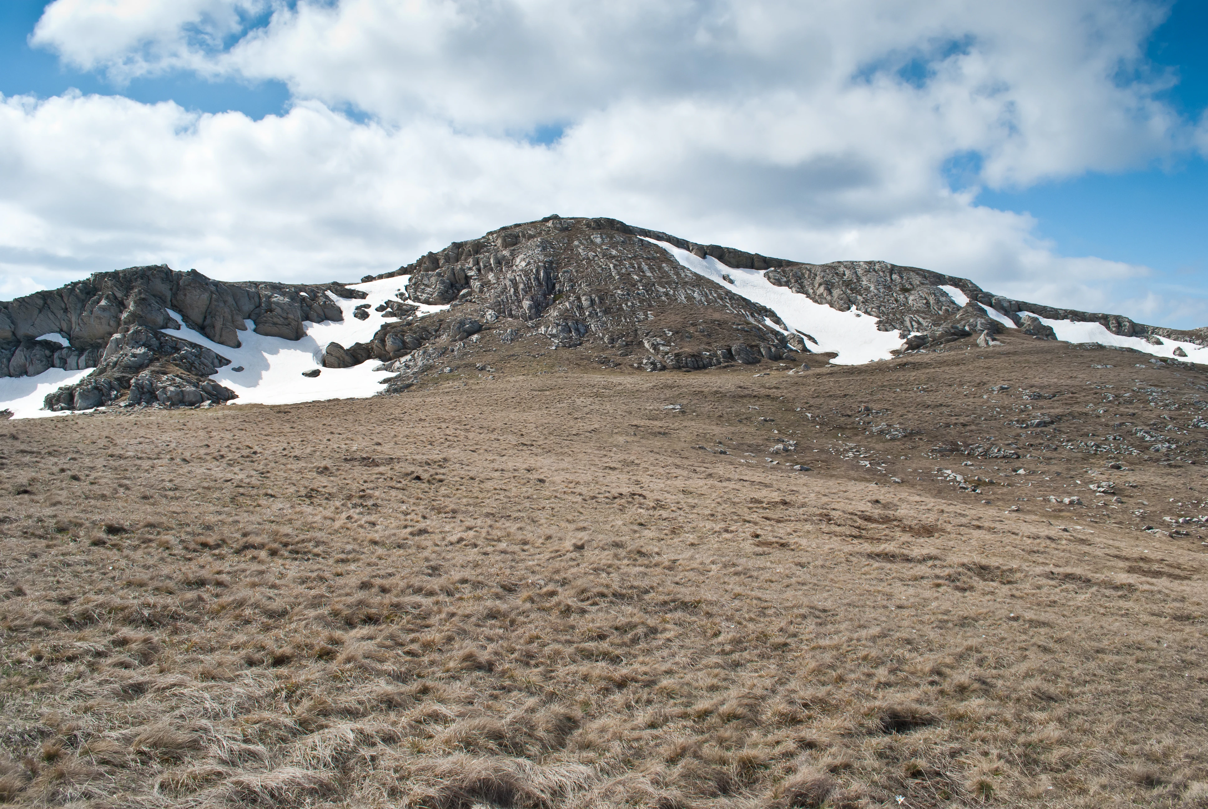 Grassy plain leading to rocky mountain slopes with snow patches under a partly cloudy sky