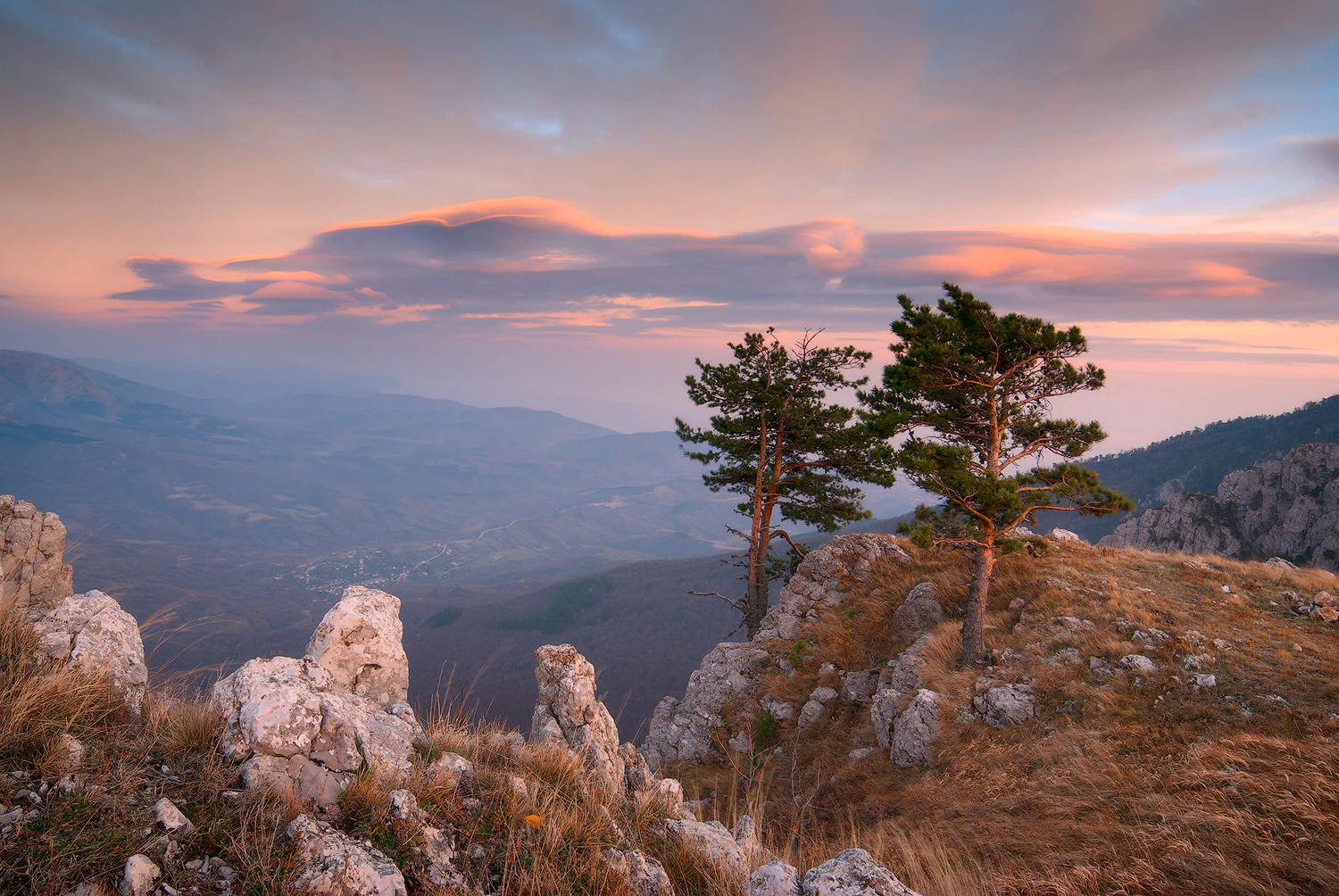Sunset sky with orange and pink hues over rocky hills and pine trees in a mountainous landscape