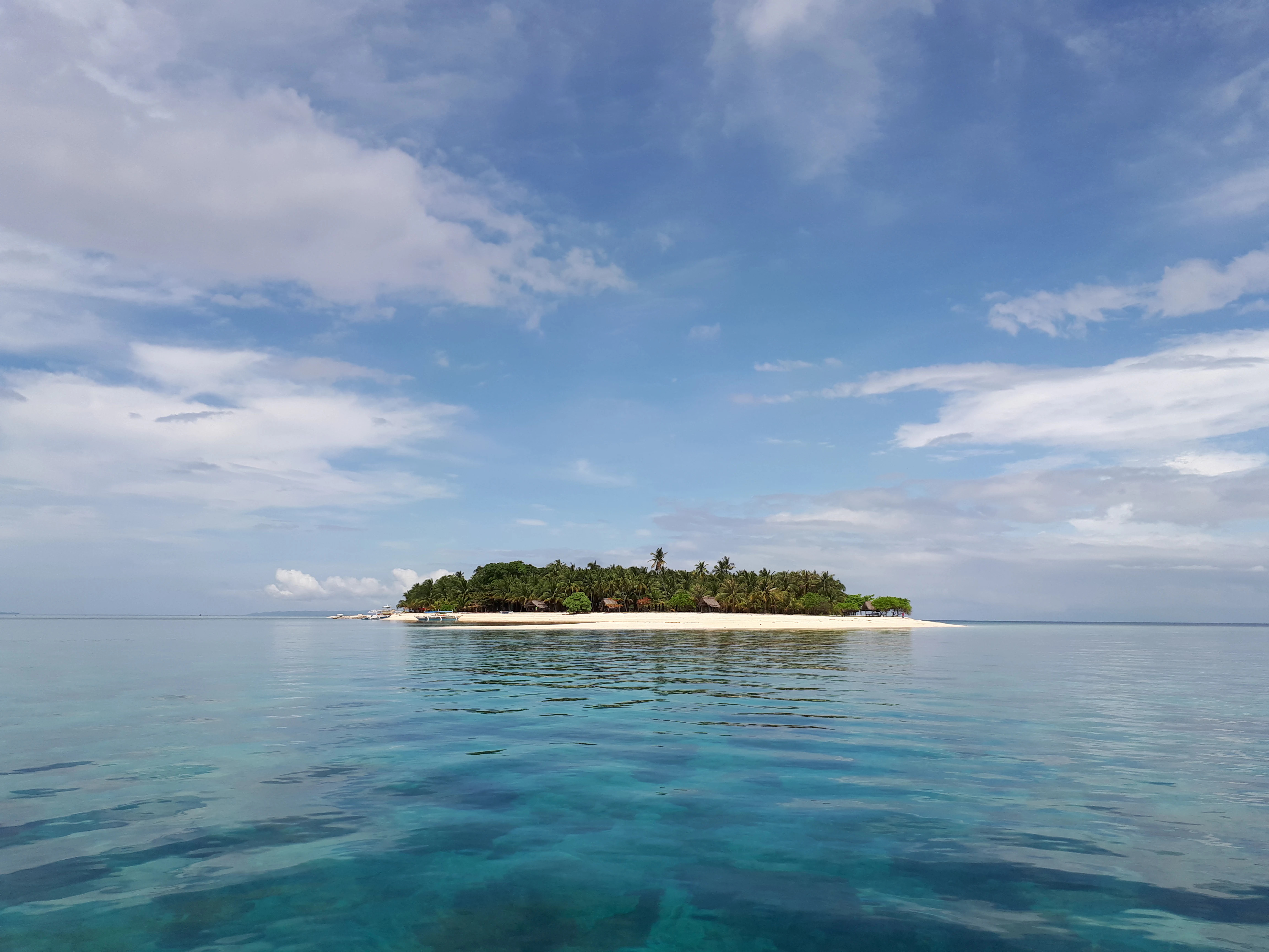 A small tropical island with a white sand beach and dense green vegetation sits in the middle of calm, clear blue water under a partly cloudy sky.