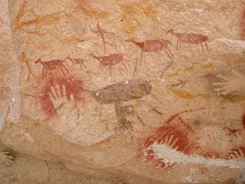 A close-up of prehistoric rock art on a textured sandstone wall, featuring red hand stencils and brown painted silhouettes of animals resembling guanacos.