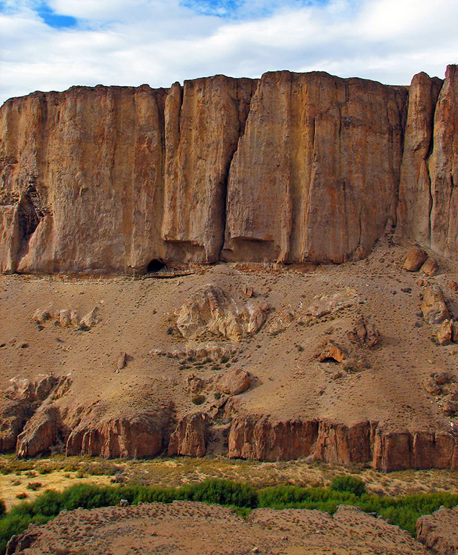 A wide shot of a tall, vertical cliff face composed of reddish-brown stone, featuring small caves and overhangs at the base, with scrubby green vegetation in the foreground.
