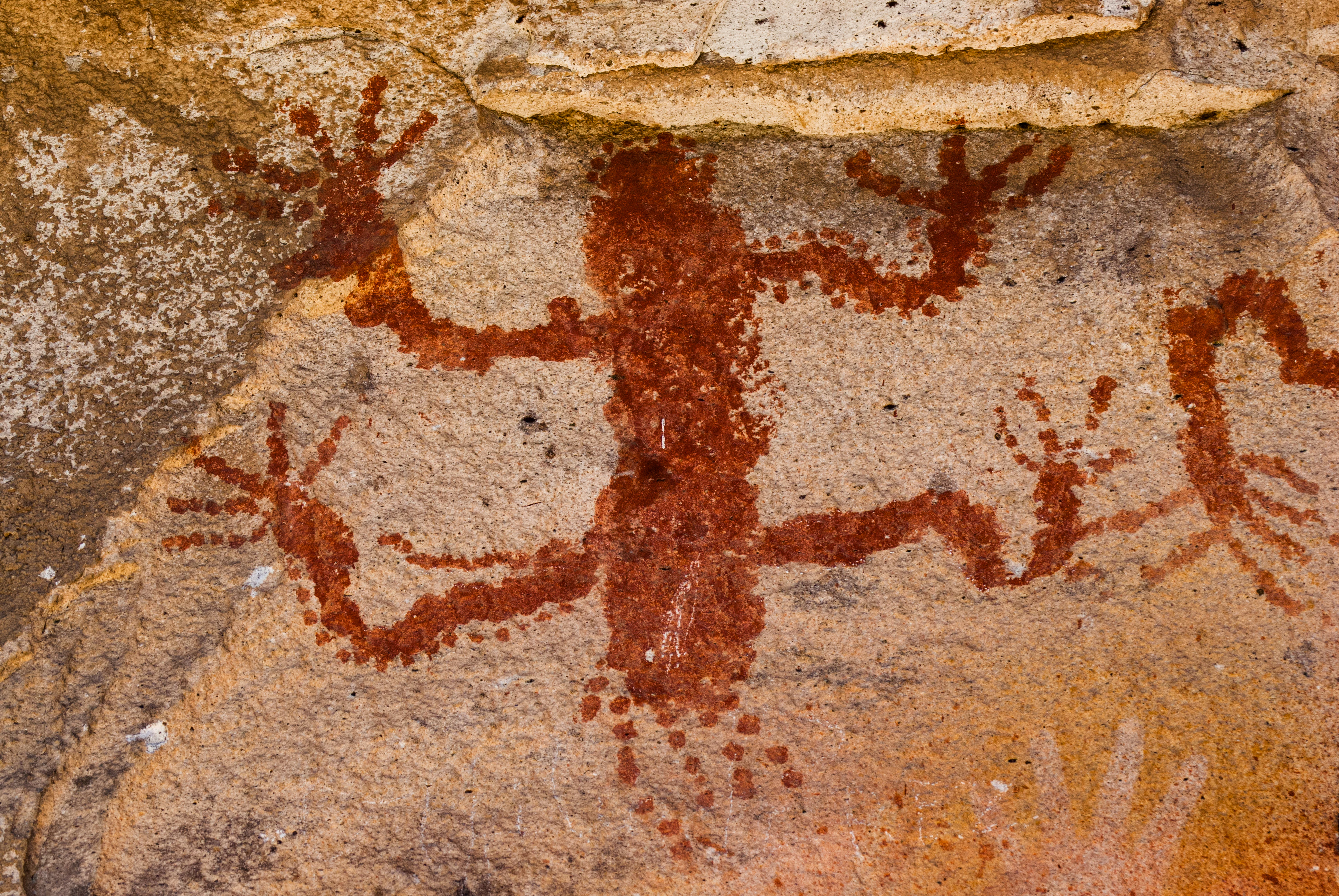 A close up view of a prehistoric red pigment painting on a textured stone wall, depicting a stylized anthropomorphic or zoomorphic figure with multiple radiating limbs.