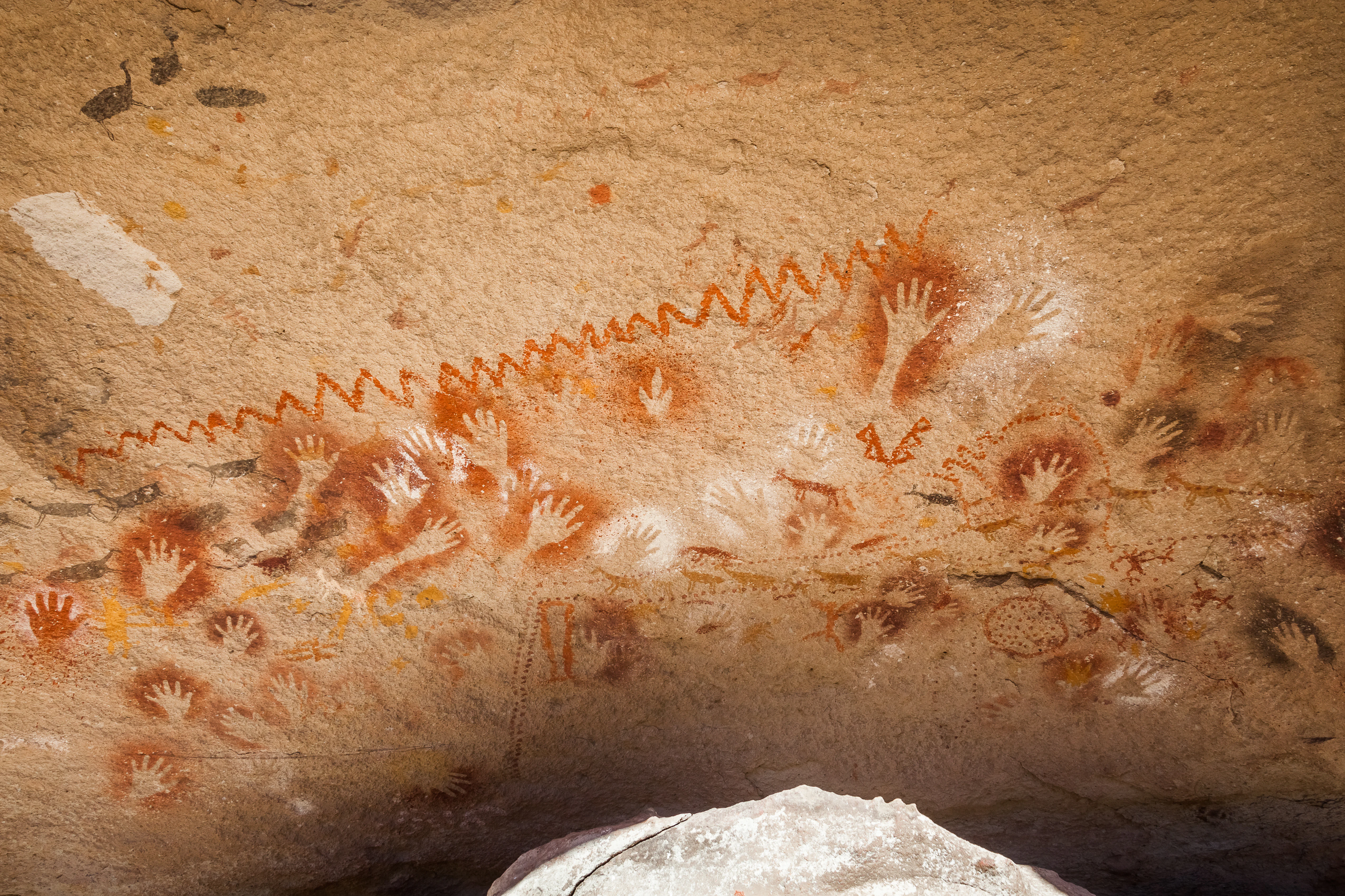 A rock wall covered in numerous ancient hand stencil paintings in red, white, and ochre, with a jagged zigzag line painted across the top.