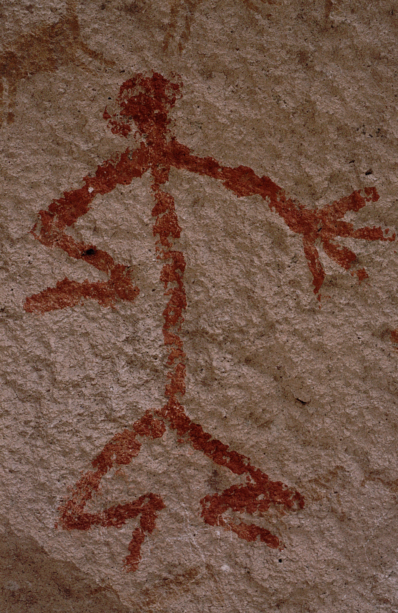 A close-up view of ancient red pigment rock art depicting a stylized human stick figure on textured rock surface.