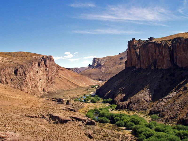 A wide view of a dry, rugged canyon with steep, rocky cliffs rising on both sides of a narrow, green, vegetated river valley.